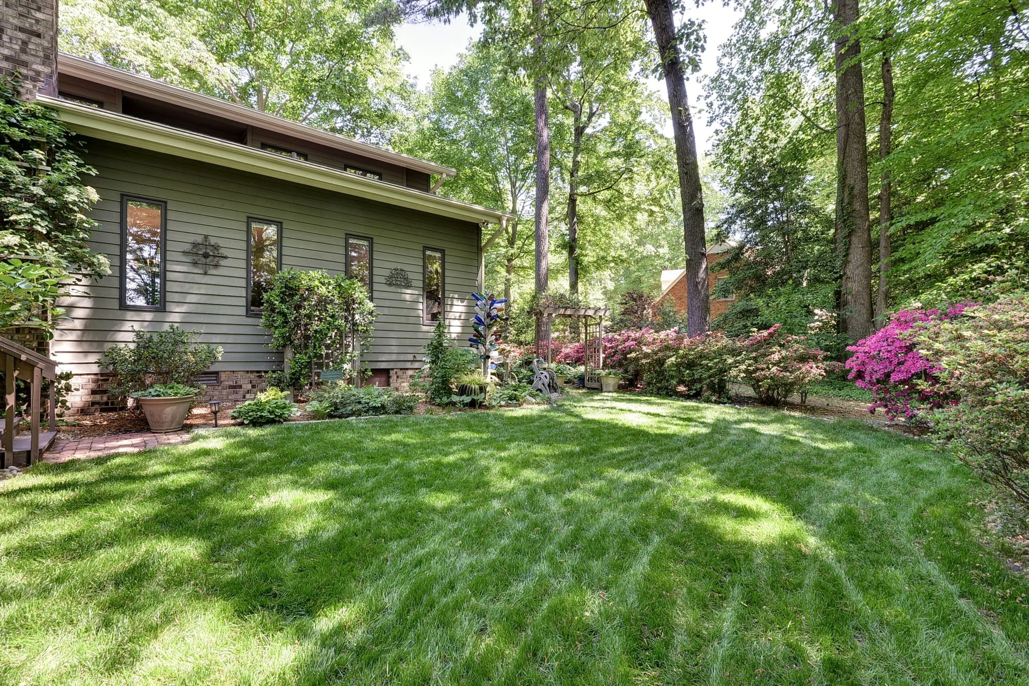 A backyard with lush green grass, various plants, trees, and colorful flowering bushes. There is a house on the left with gray siding and several windows, along with garden decorations and outdoor furniture.
