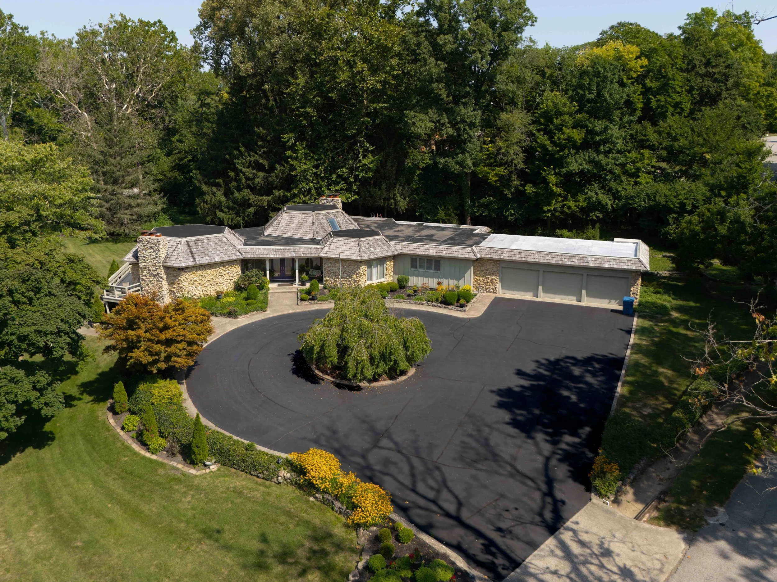 A large house with stone exterior, a driveway, and a three-car garage, surrounded by green trees and landscaped yard.