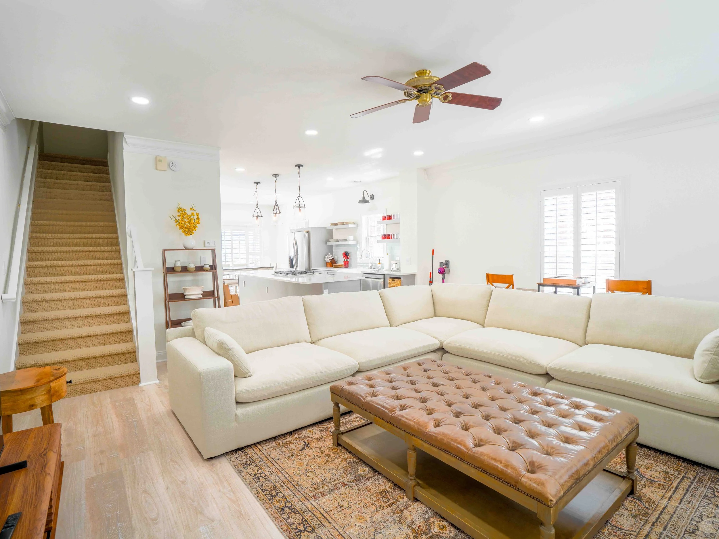 Bright living room with a large off-white sectional sofa, a brown leather tufted coffee table, and a patterned area rug. In the background, there's a white open-concept kitchen with pendant lighting, stainless steel appliances, and a dining area with wooden chairs. A staircase with beige carpet leads upstairs, and windows with white shutters let in natural light.