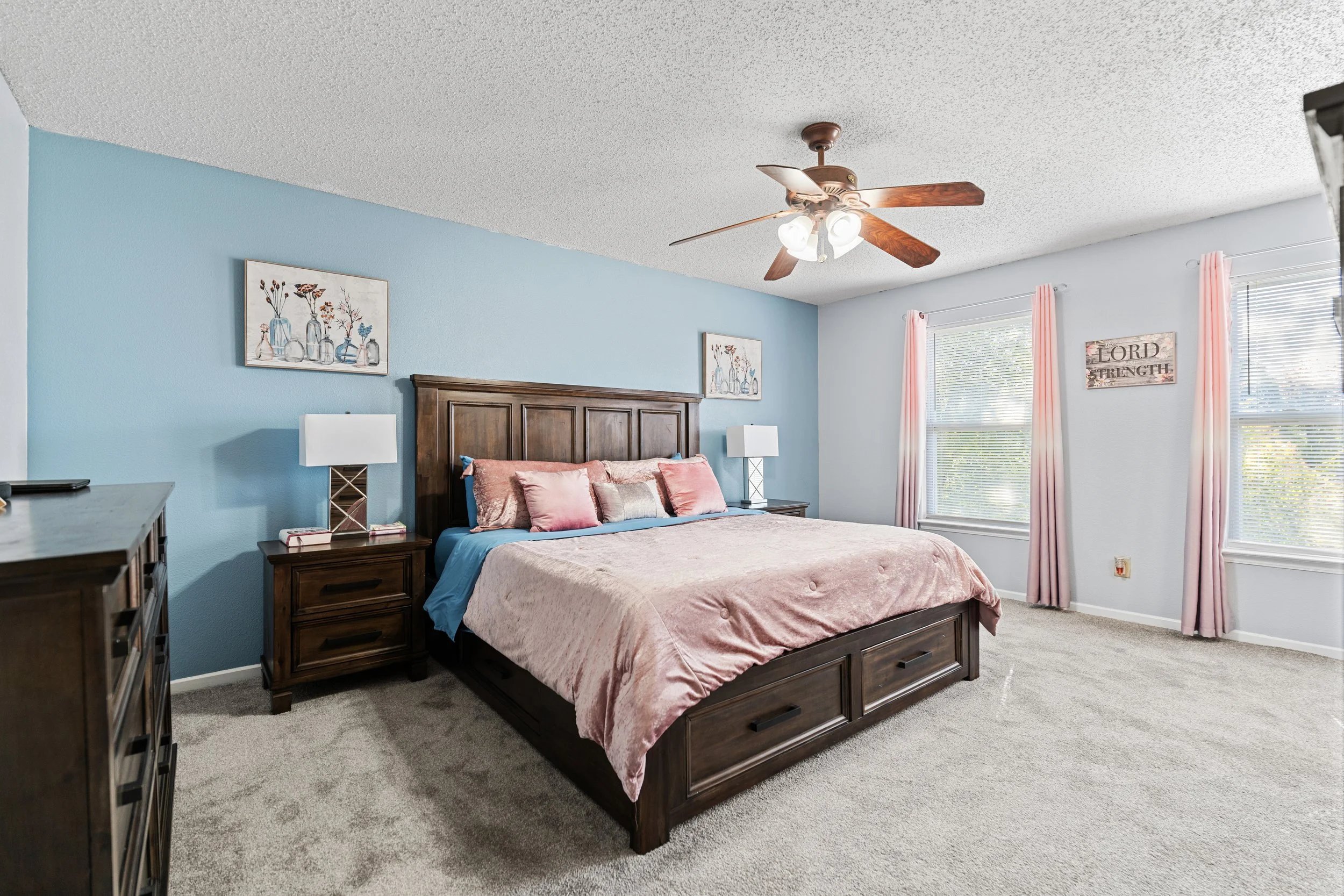 A bedroom with a large bed, pink and beige bedding, dark wood furniture, two windows with pink curtains, wall art, a ceiling fan, and a light blue accent wall.
