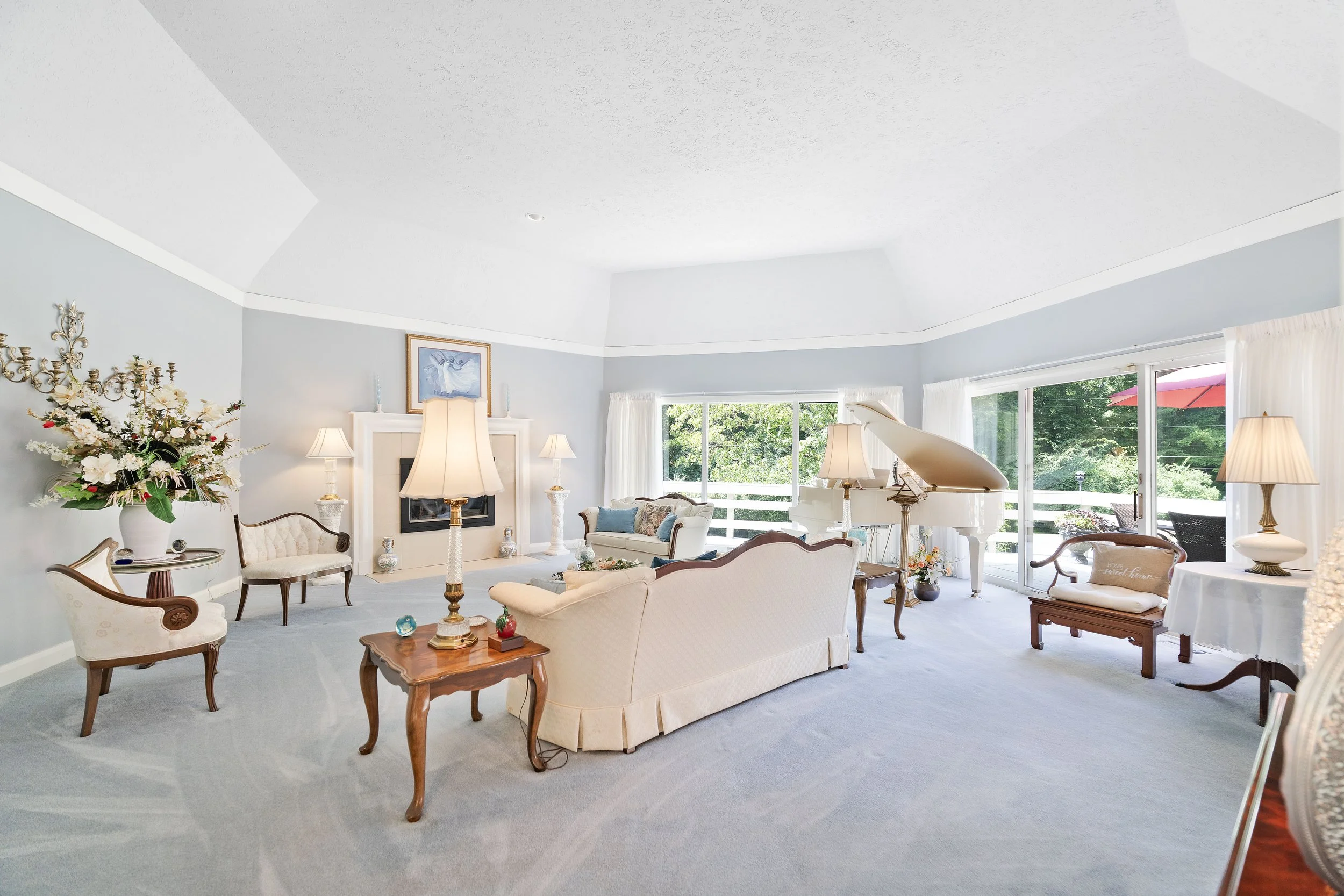Bright living room with white sofa, vintage chairs, grand piano, floral arrangements, large windows, and outdoor view