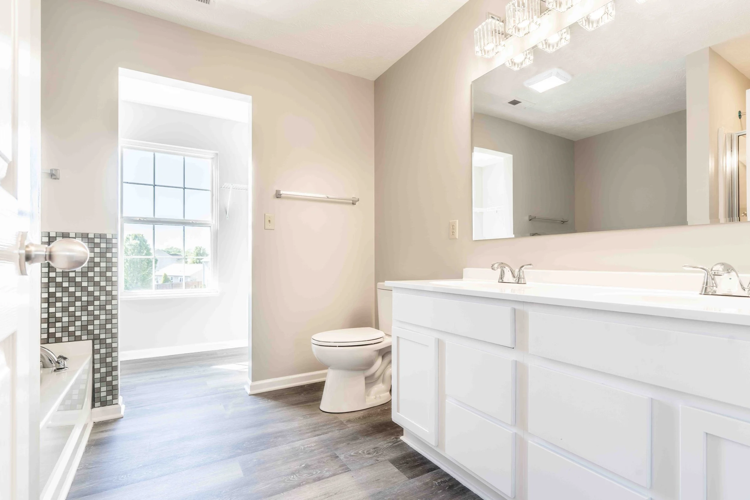 Clean bathroom with a double white vanity, large mirror, toilet, and window with sunlight coming in.