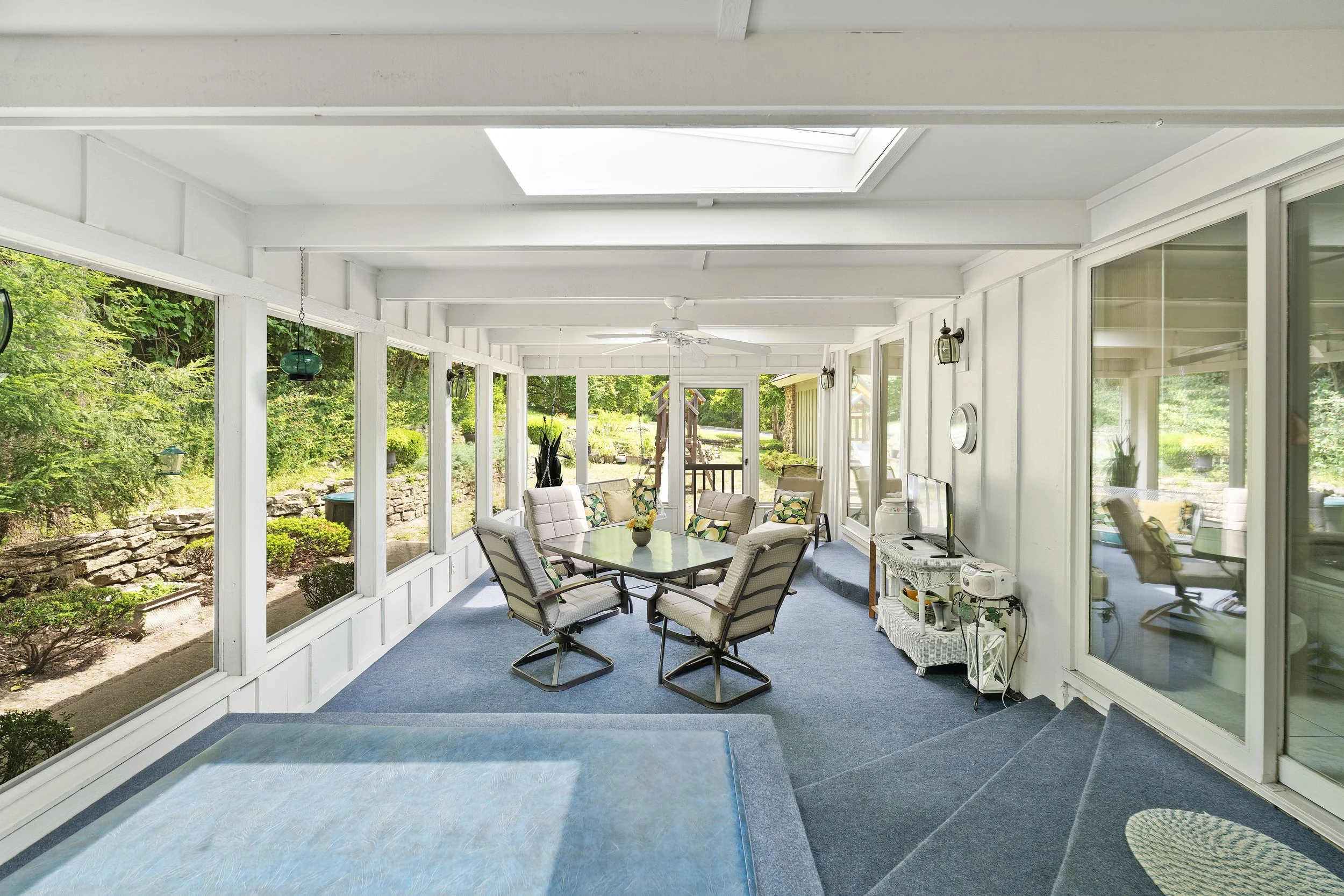 Sunroom with blue carpet, glass walls showing green outdoor scenery, dining table with six chairs, corner with small white shelf, and sliding glass door leading outside.