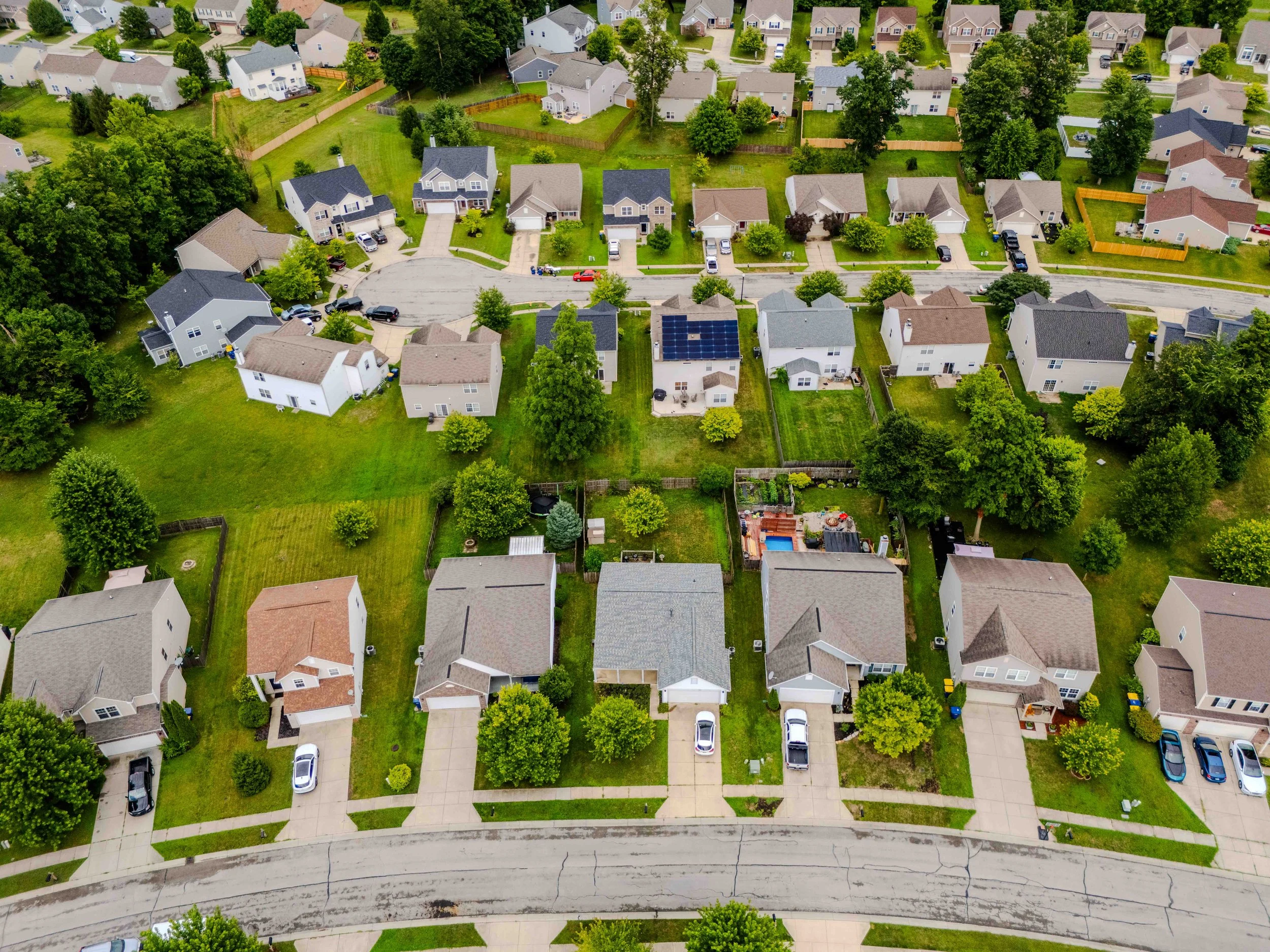 Aerial view of a suburban neighborhood with houses, green lawns, trees, and cars parked in driveways and on the street.
