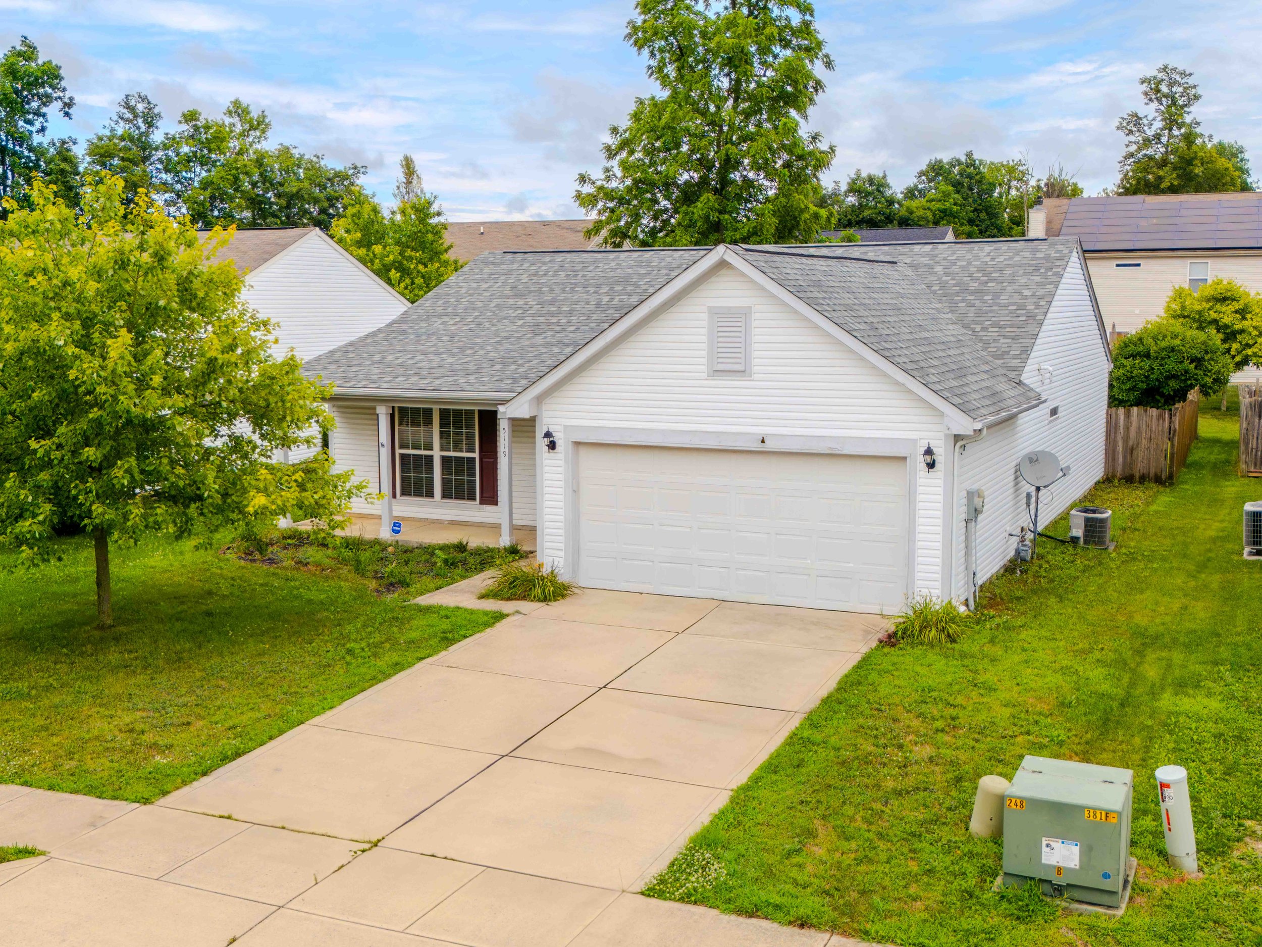 A white, single-family house with a two-car garage, a front porch, and a sloped shingle roof. There are green trees and grass surrounding the house, with a concrete driveway leading to the garage. Utility meters and satellite dishes are visible on the side of the house.