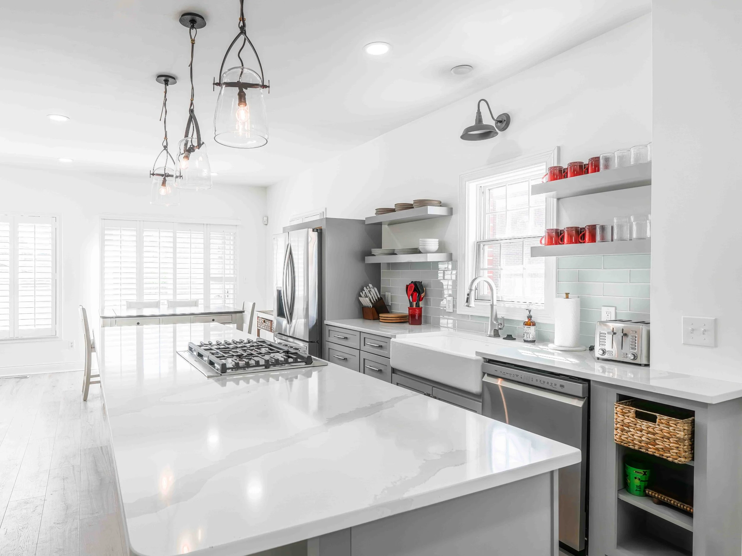 Modern kitchen with white countertops, gray cabinets, open shelves with red and clear glasses, stainless steel appliances, a farmhouse sink, and pendant lighting fixtures.