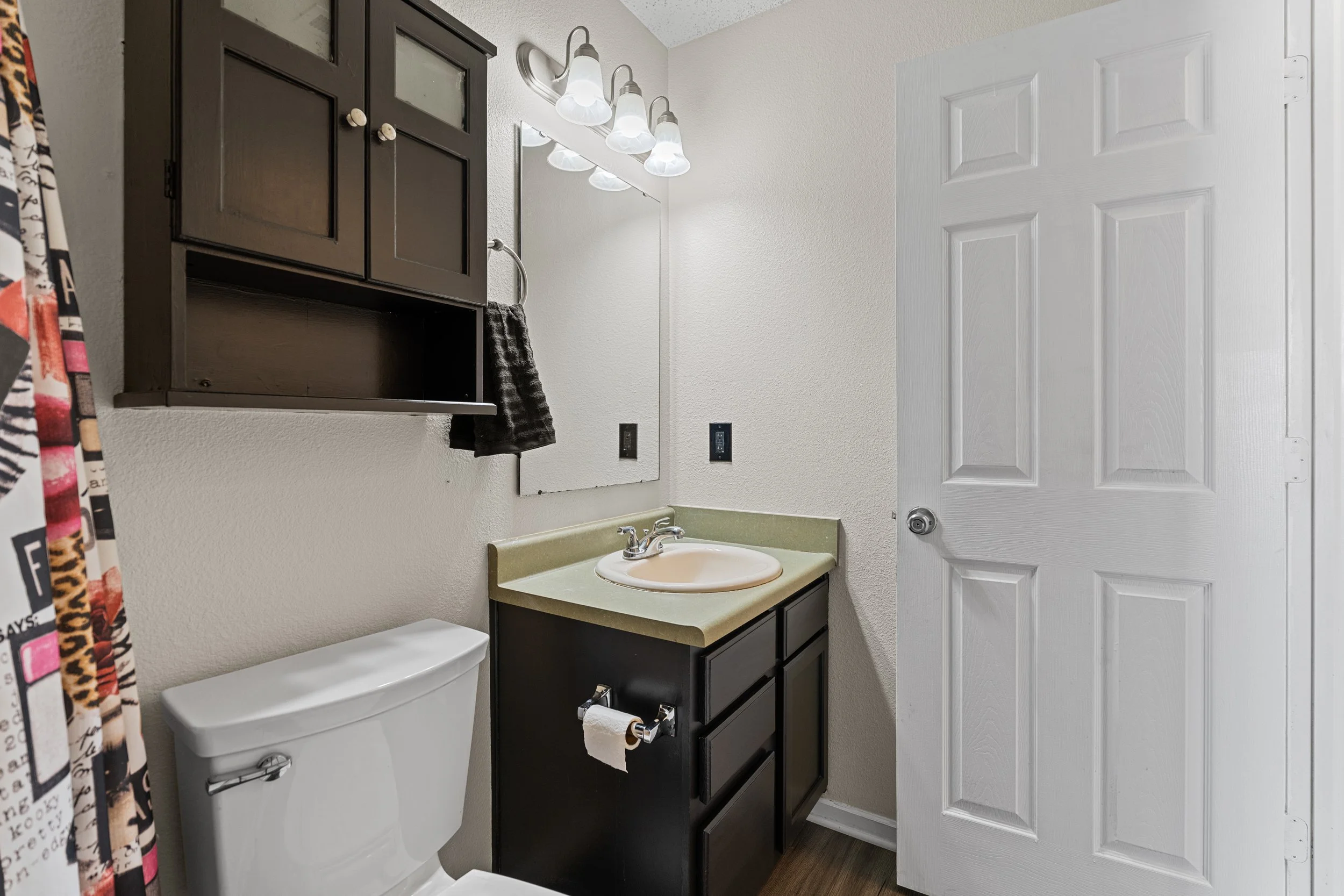 Small bathroom with a white toilet, black cabinet with beige sink, wall-mounted mirror, black wall cabinet, and light fixture with four bulbs. White door partially open, patterned shower curtain on the left, and beige textured walls.