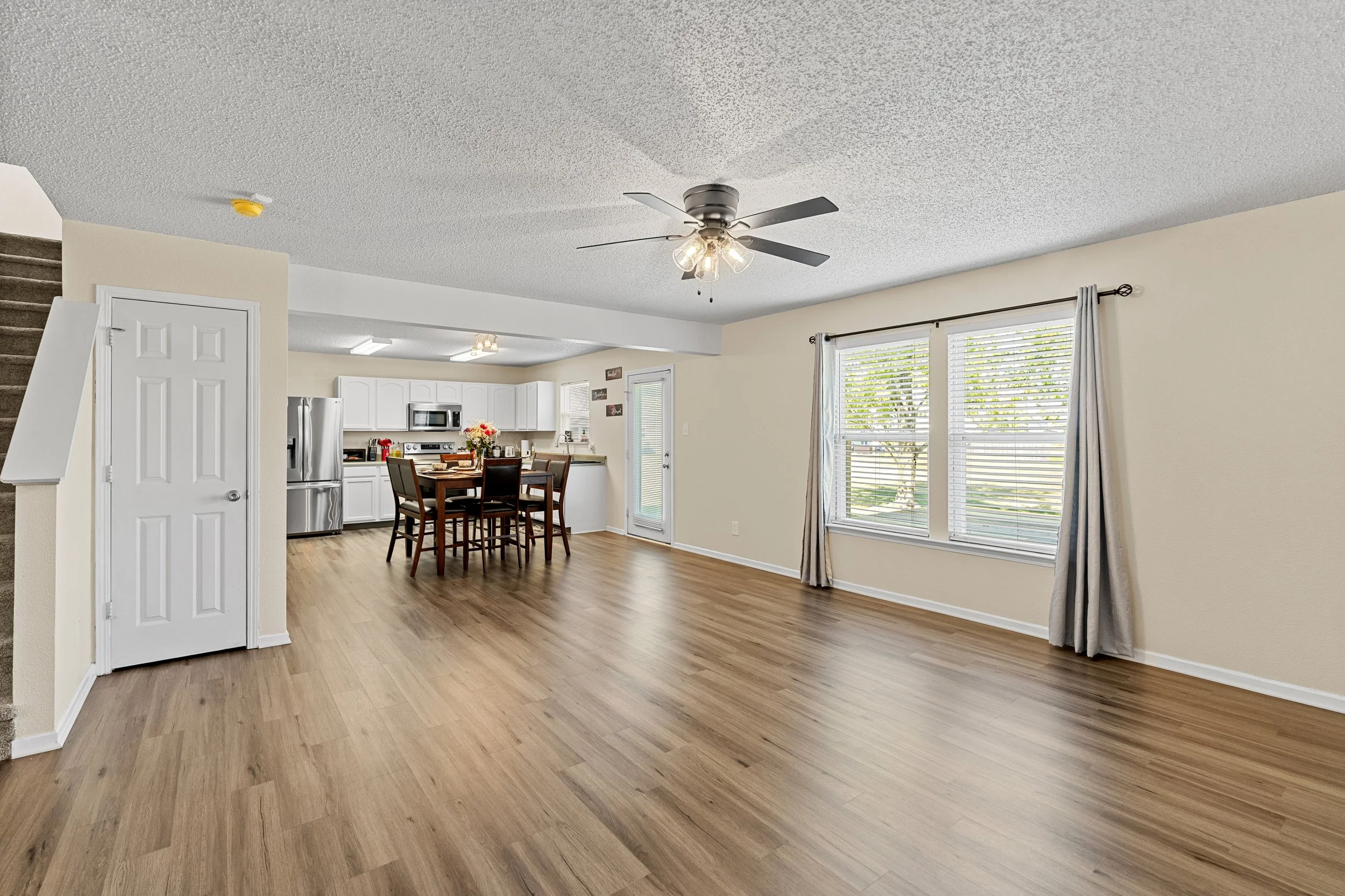 Empty living room and dining area with hardwood floors, large windows with blinds and curtains, ceiling fan with lights, adjacent kitchen with stainless steel appliances and white cabinets.
