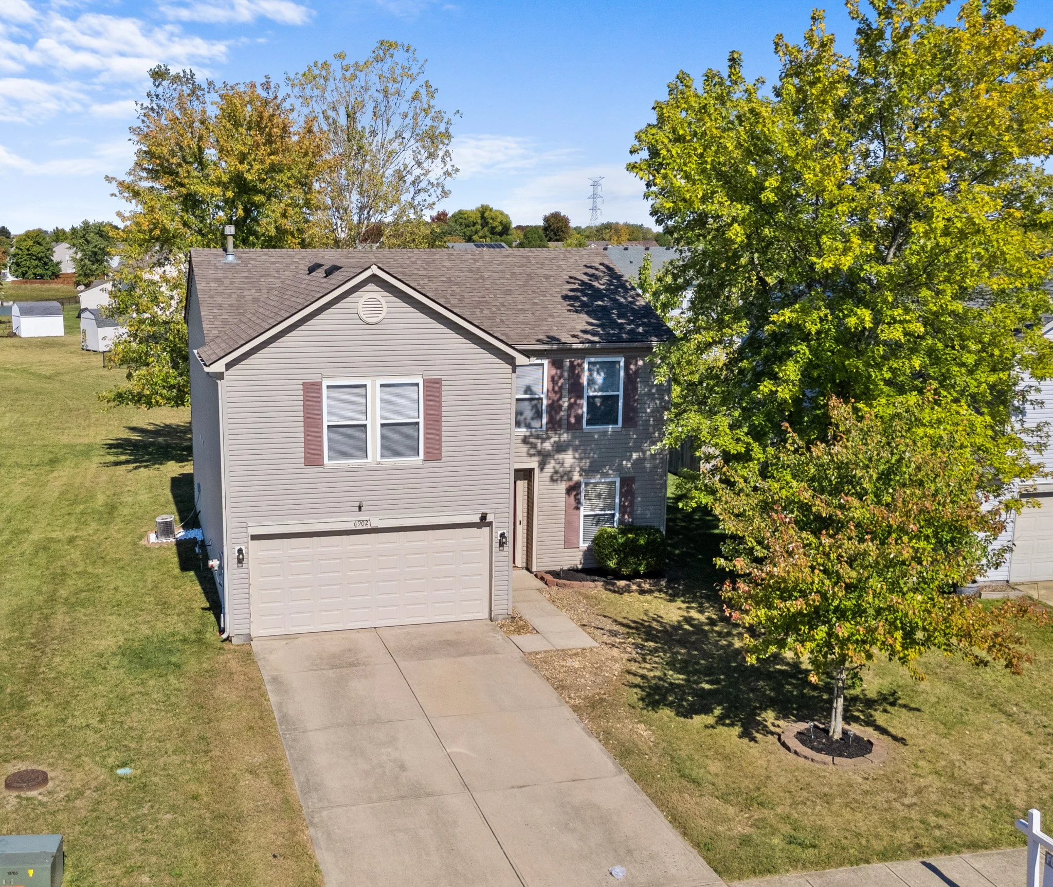 A two-story house with a beige exterior, a front garage, and a concrete driveway. There are two large trees, one on each side of the house, with green and yellow leaves. The house has three visible windows with brown shutters, and a small entrance porch with a door. The lawn is green with some patches of dirt, and neighboring houses are visible in the background under a blue sky with some clouds.