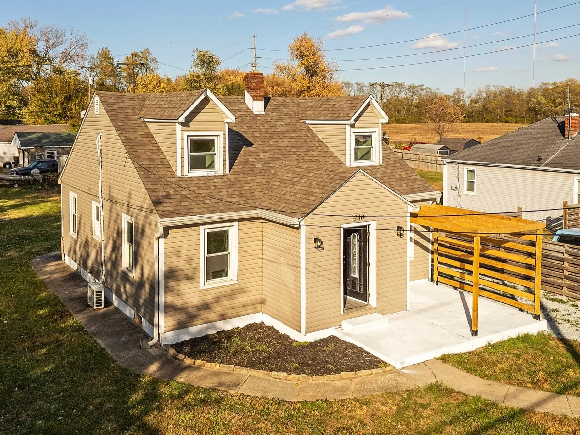 A beige house with a brown shingle roof, two small dormer windows, and a front door with an overhang, located in a suburban neighborhood.