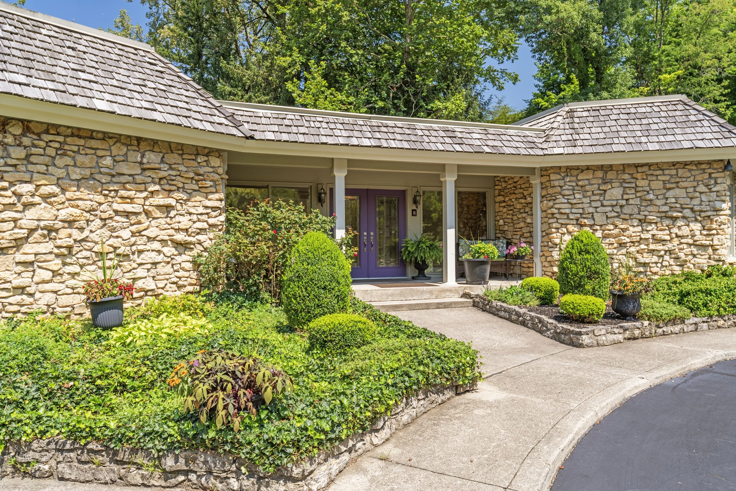 Front view of a house with stone exterior walls, purple front door, and a small porch with plants and flowers in pots, surrounded by a well-maintained garden and greenery.