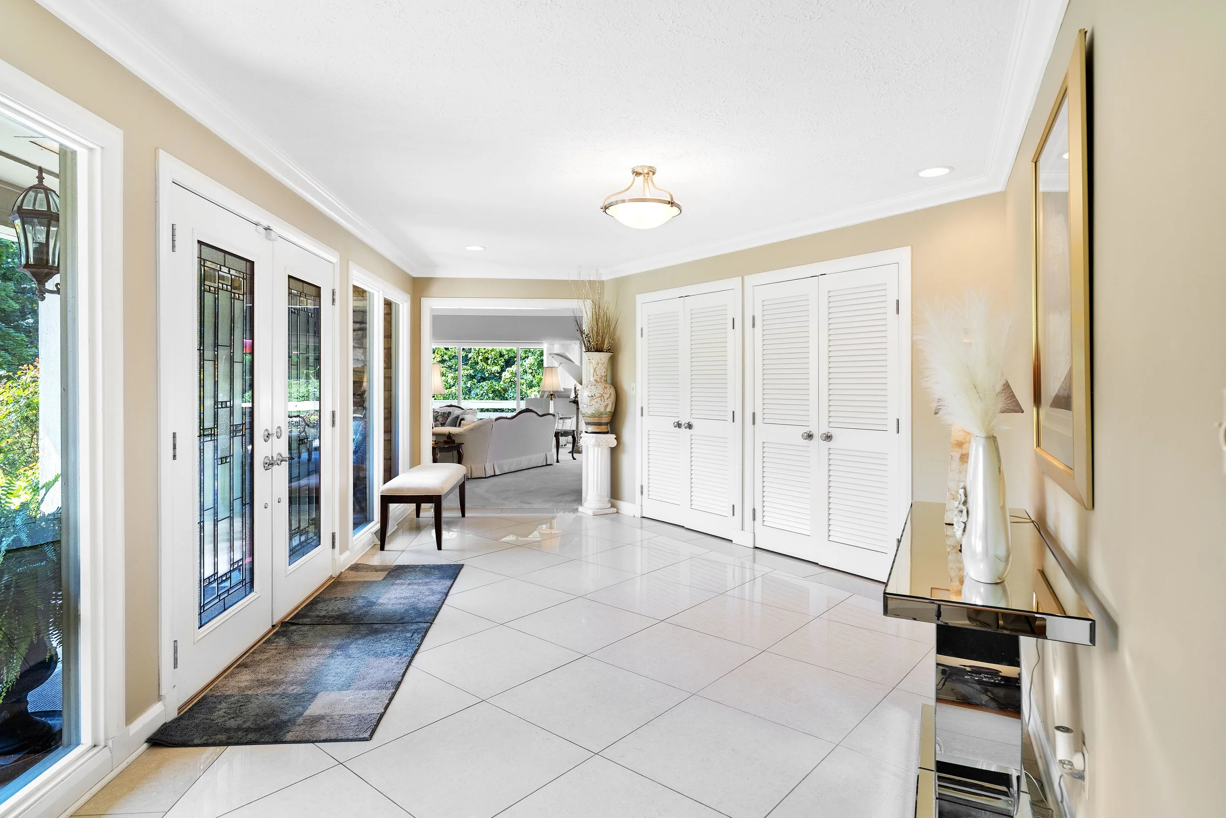 Bright foyer with tiled floor, double doors with stained glass, and a bench near the entrance. Opens into a living room with large window, sofas, and plants.