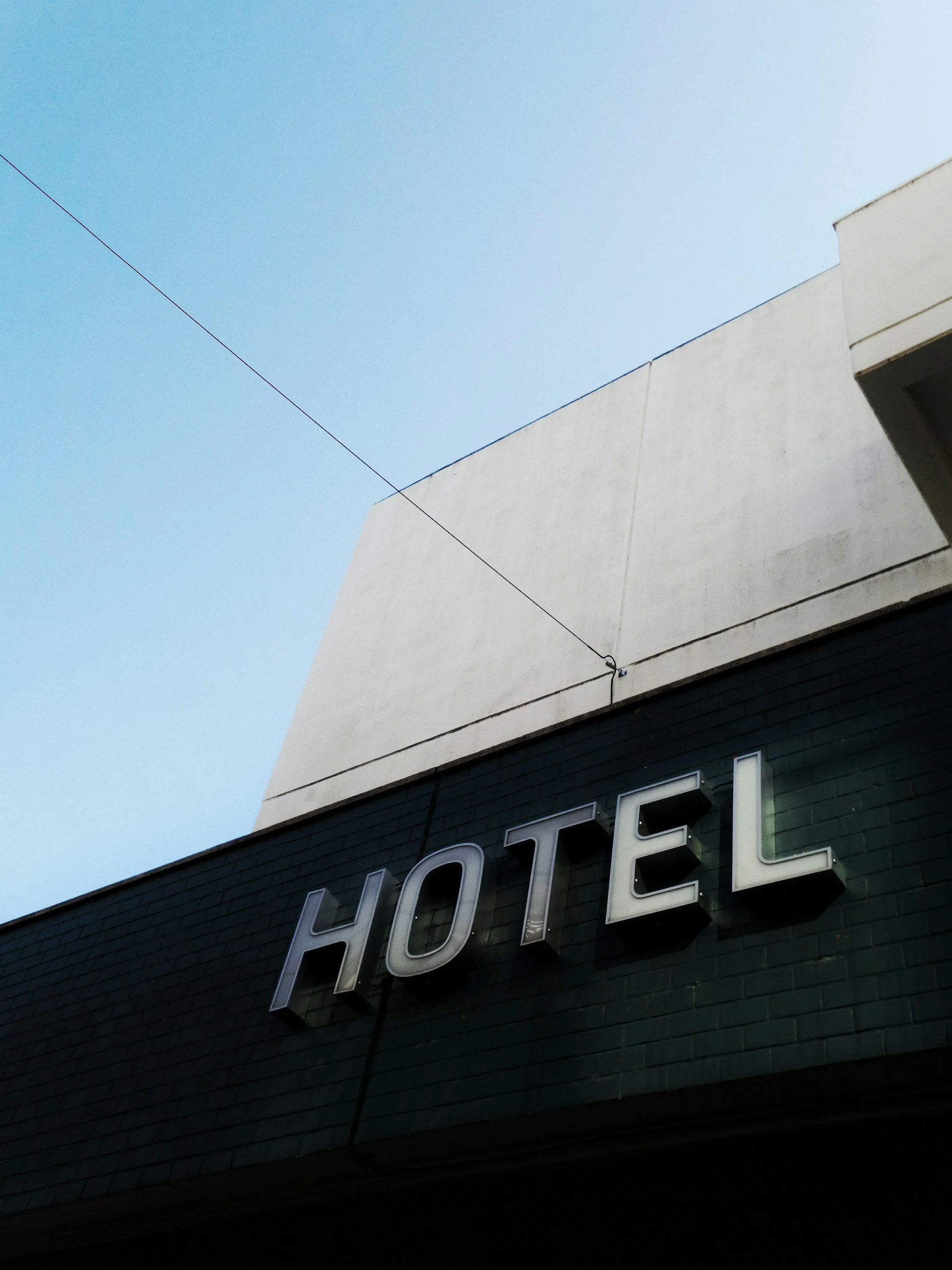 Close-up of a black building partial sign reading 'HOTEL' with a white geometric building structure in the background against a clear blue sky.