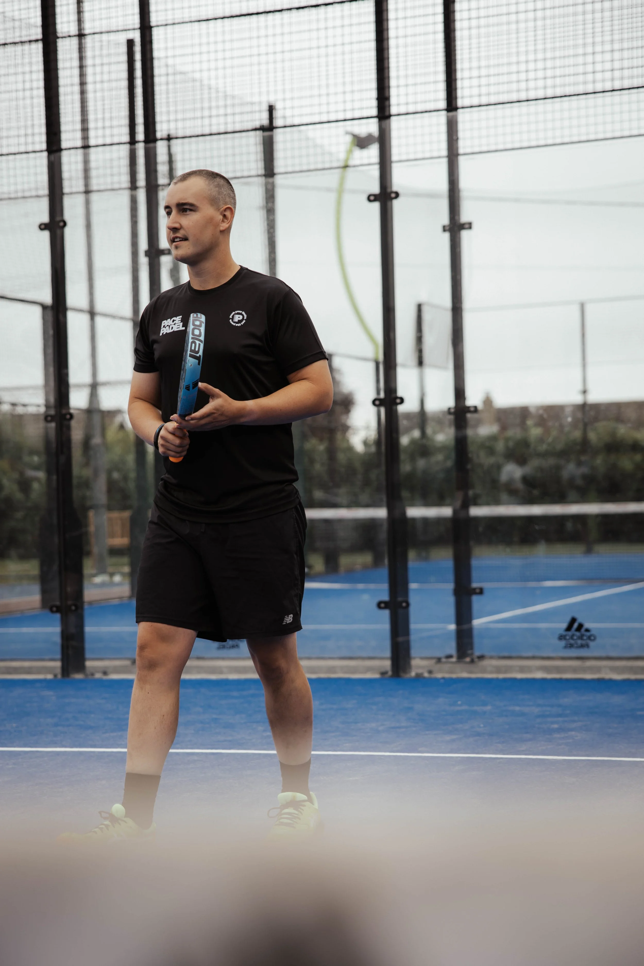 A man standing on a blue padel court, holding a padel racket, with a netted enclosure and tennis courts in the background.