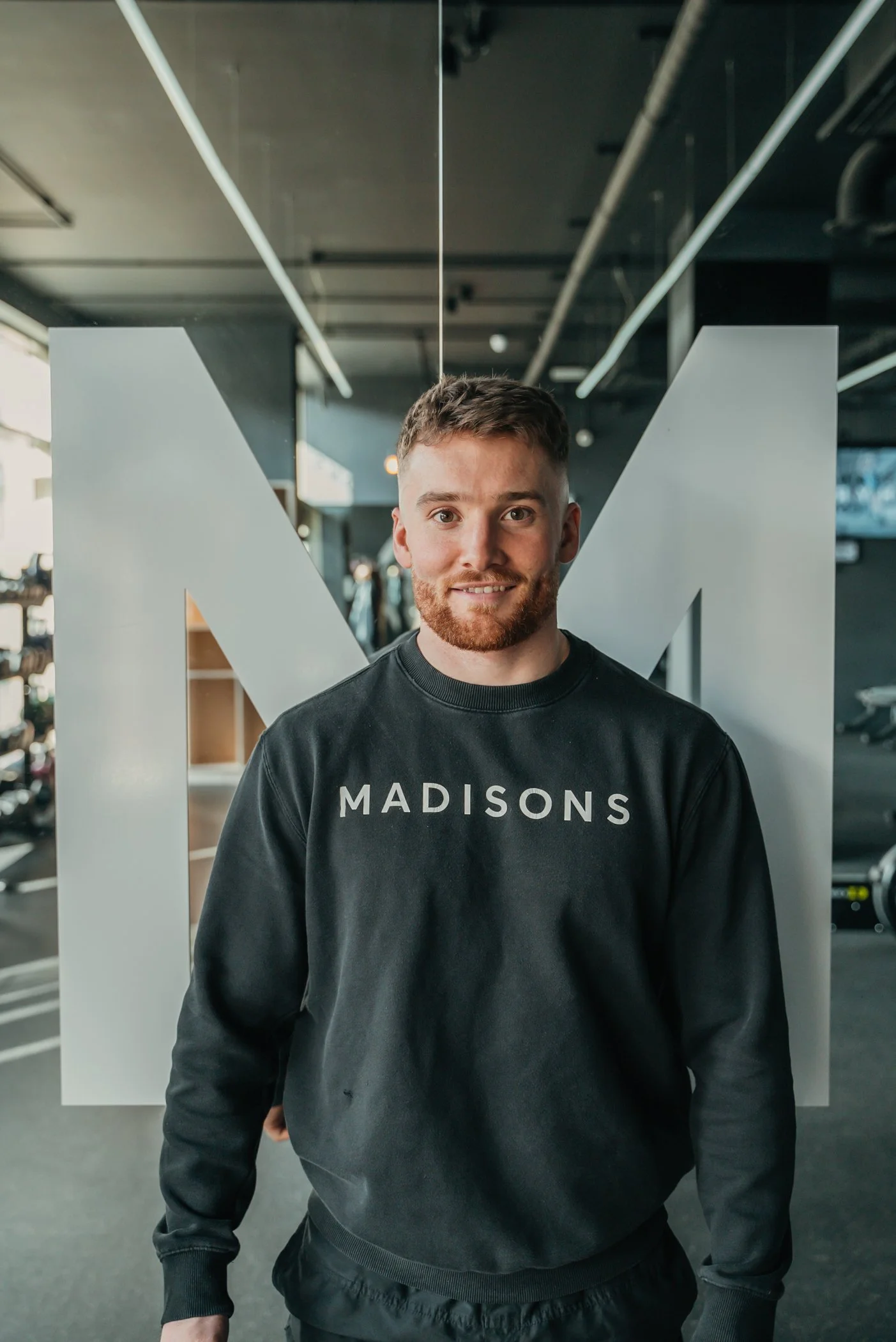 A young man with short brown hair and a beard, wearing a black sweatshirt that says 'MADISONS,' standing inside a gym with modern equipment and large white letter 'M' in the background.