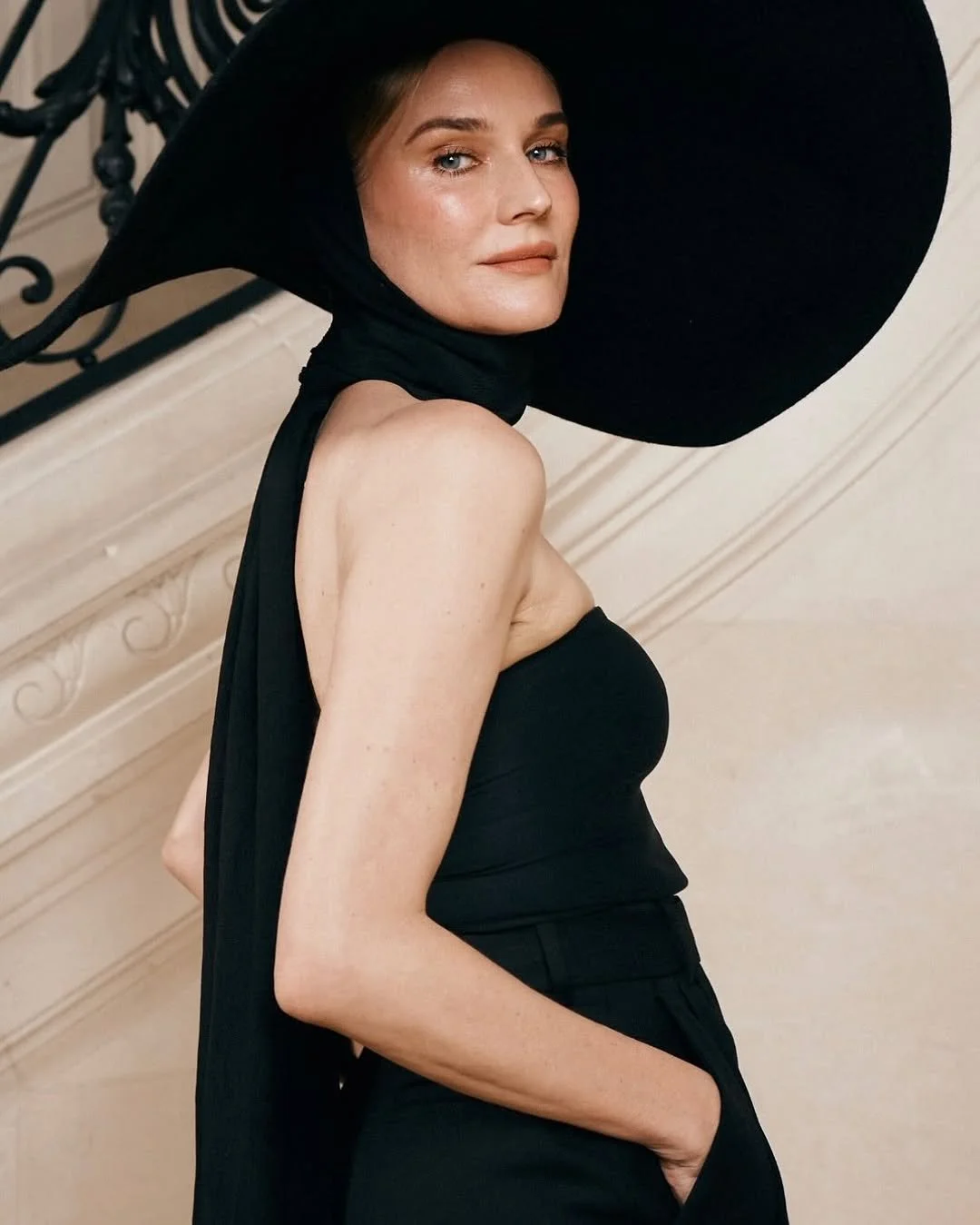 Femme portant un grand chapeau noir, tenue noire, pose près d'une rampe en fer forgé, intérieur élégant.