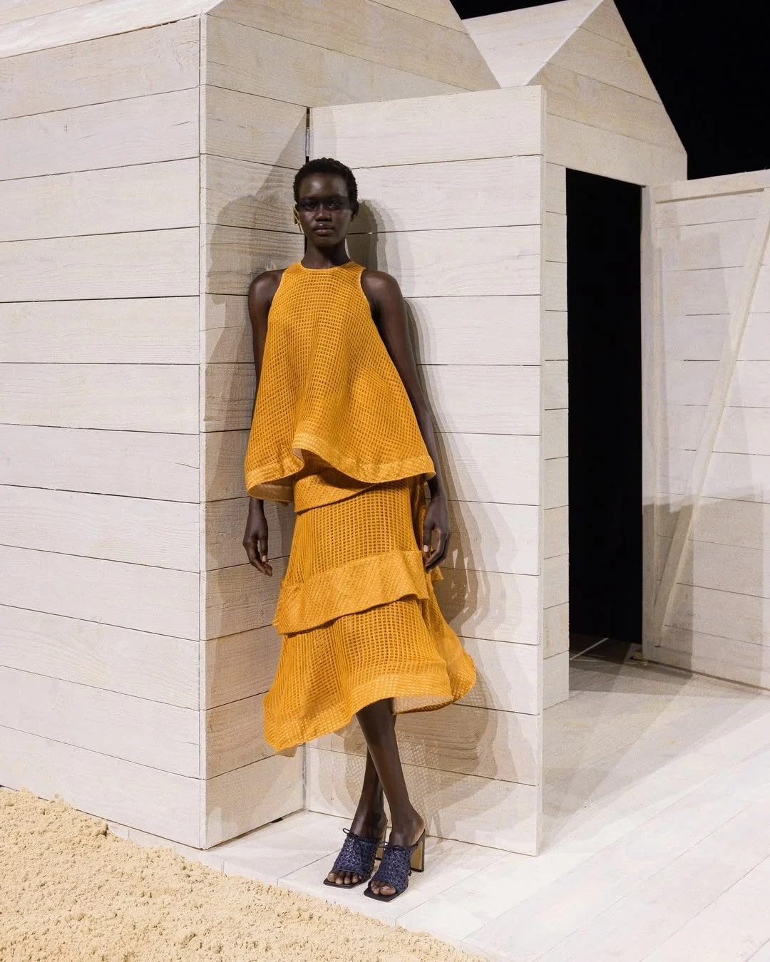 Une femme portant une robe longue orange avec un haut ample et une jupe à plusieurs couches, se tient contre un mur en bois clair, dans un décor minimaliste.