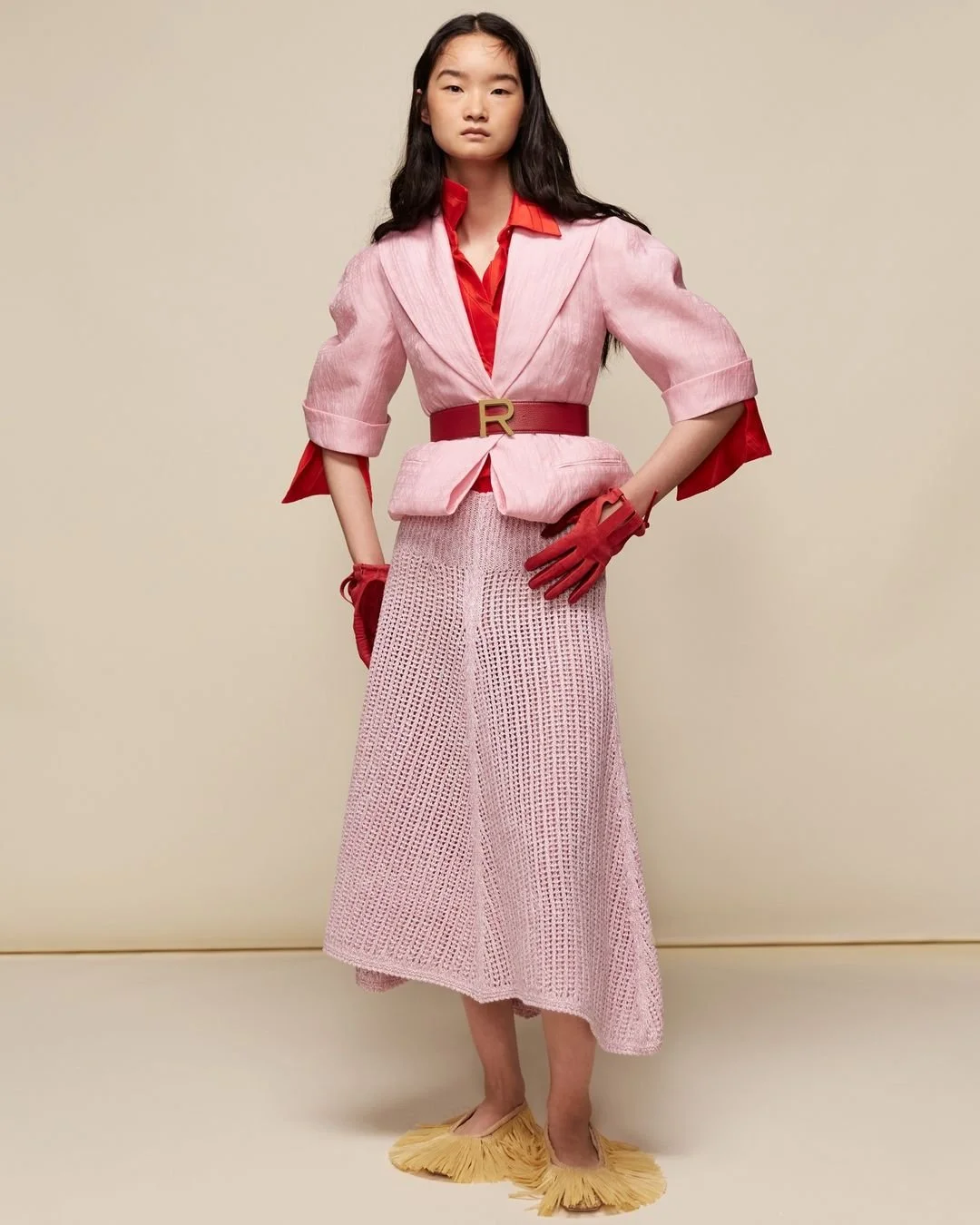 Une jeune femme porte une tenue de mode en couleurs rose et rouge, avec des gants rouges, un long jupon rose en crochet, des chaussures jaunes à franges et une ceinture rouge avec un bouton en forme de lettre R.