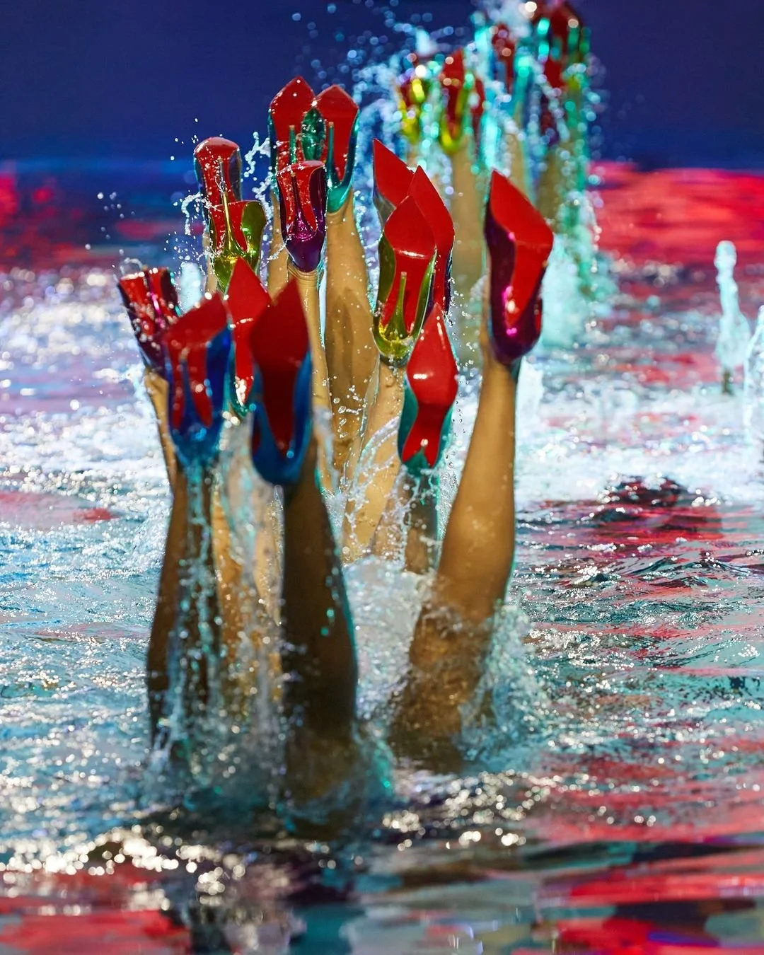 Des jambes de nageuses portant des chaussures à talons rouges, vue de dessous, en train de plonger ou de se relever dans une piscine, avec de l'eau éclaboussée autour.