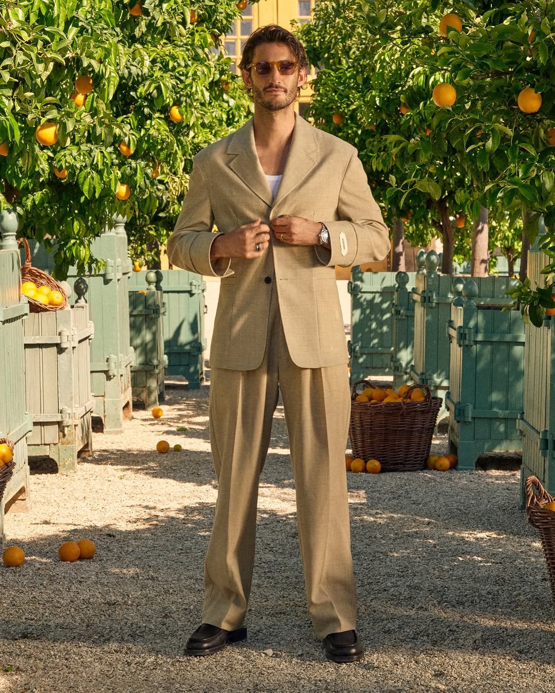 Un homme en costume beige se tient dans un verger avec des orangers. Il porte des lunettes de soleil, une montre, et ajusté son blazer.