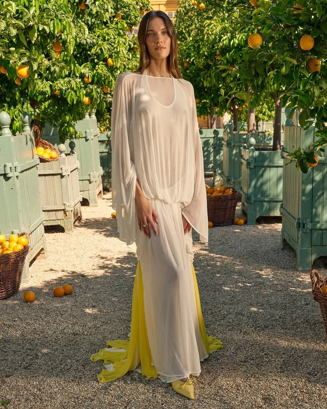 Femme en tenue blanche et jaune dans un verger d'orangers avec des paniers d'oranges.