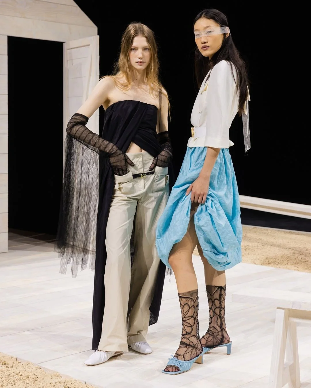 Deux femmes posent avec des vêtements de mode sur un plateau de studio. La première porte un haut noir, un pantalon beige et des gants noirs en filet. La seconde porte une veste blanche, une jupe bleu clair nouée, des chaussures à talons bleues, des bas en résille noire avec un motif et un cache-œil transparent.