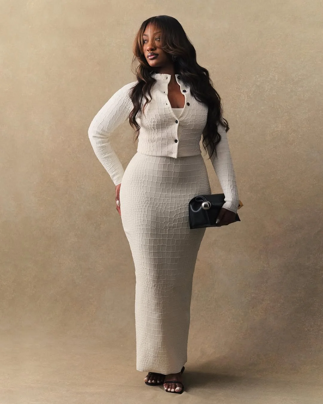 Femme portant une robe longue beige, un cardigan assorti, tenant un petit sac noir, debout contre un fond neutre.