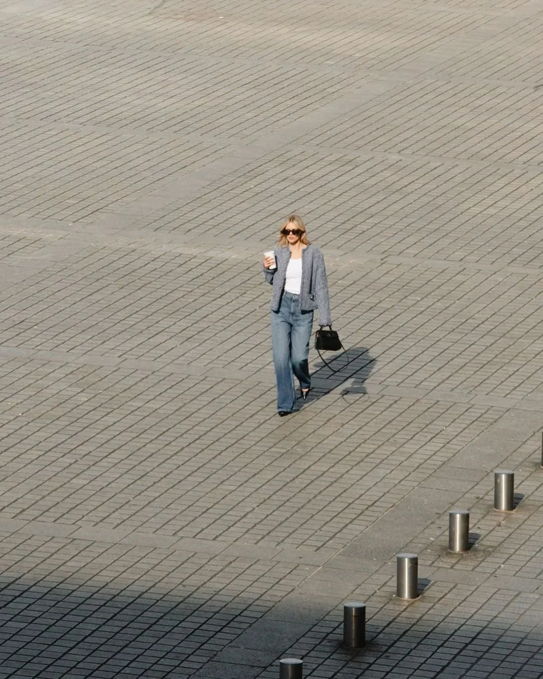 Femme marchant dans une grande place pavée, portant un pantalon bleu, une veste gris, sunglasses, tenant une boisson, avec un sac noir.