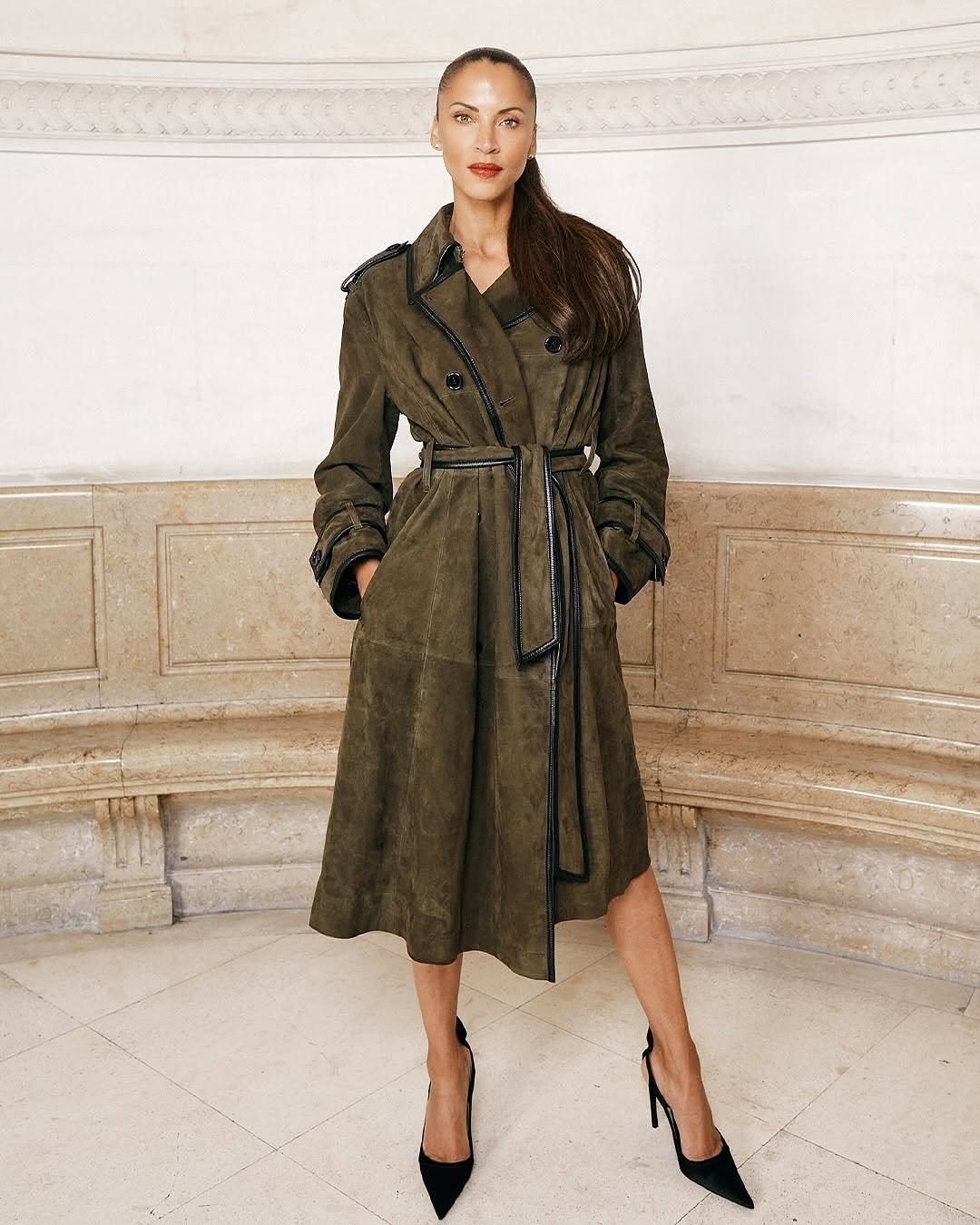 Femme portant un trench-coat olive foncé avec chaussures à talons noirs, dans un intérieur en pierre classique.