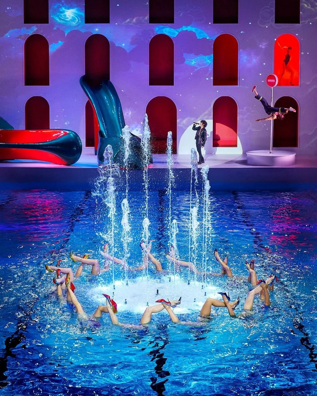 Spectacle aquatique avec des nageurs portant des jupes musicales, une personne chantant, et une acrobate volant autour d'un mât, dans un décor coloré avec un bâtiment fantaisiste et des jets d'eau.
