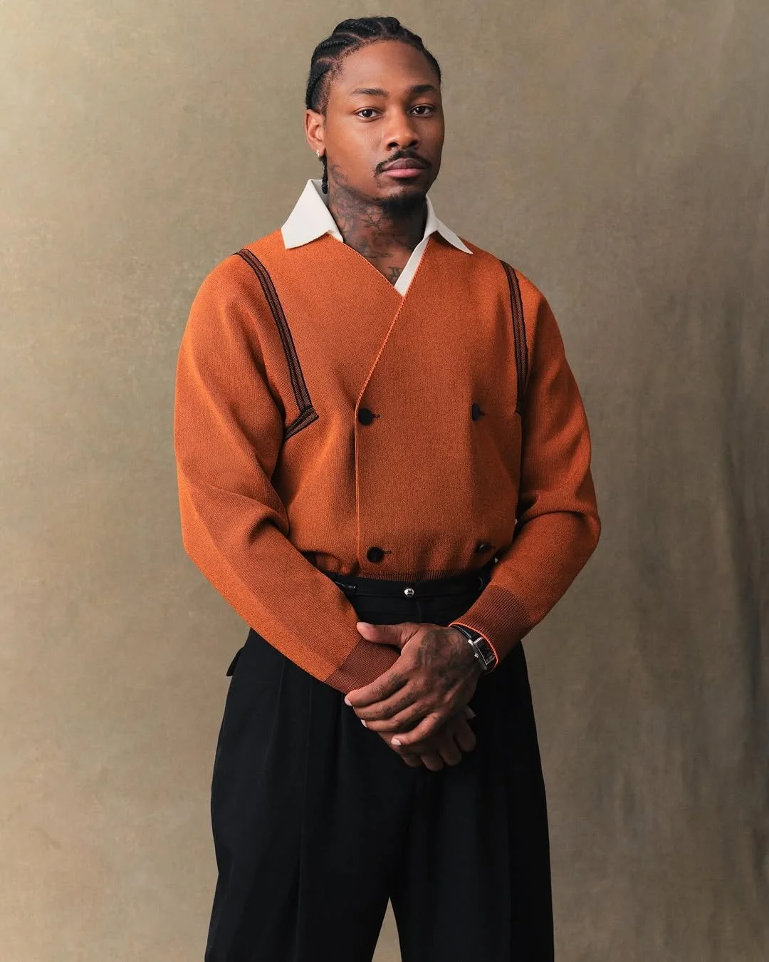 Homme avec dreadlocks, portant un pull marron avec col blanc et pantalon noir, posé contre un fond beige.