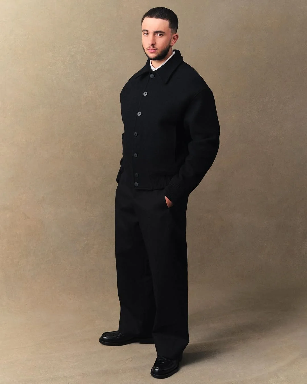 Jeune homme portant un manteau noir, chemise blanche et pantalon noir, debout devant un fond beige.