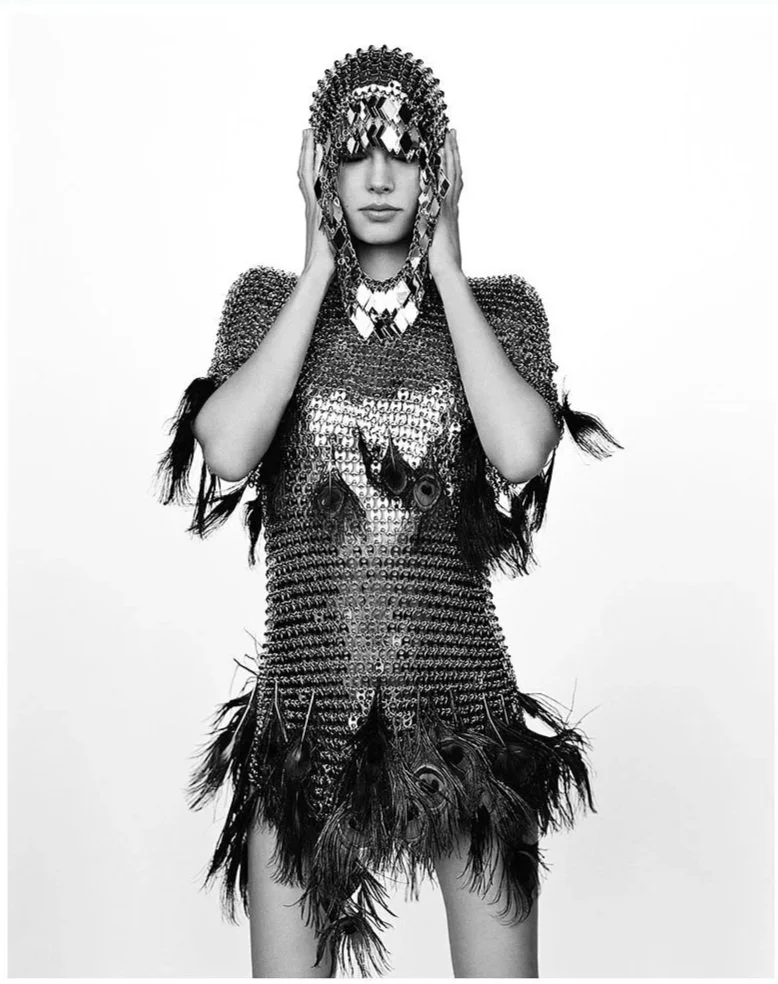 Une femme portant un vêtement et un accessoire en sequins et plumes, tenant un masque en miroir devant son visage.