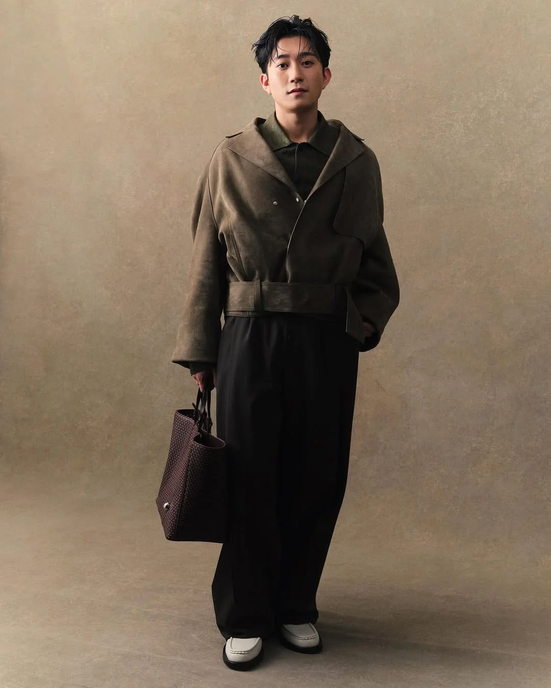 Un homme debout contre un fond beige, portant un manteau marron, un pantalon noir large, des chaussures blanches, et tenant un sac à main marron