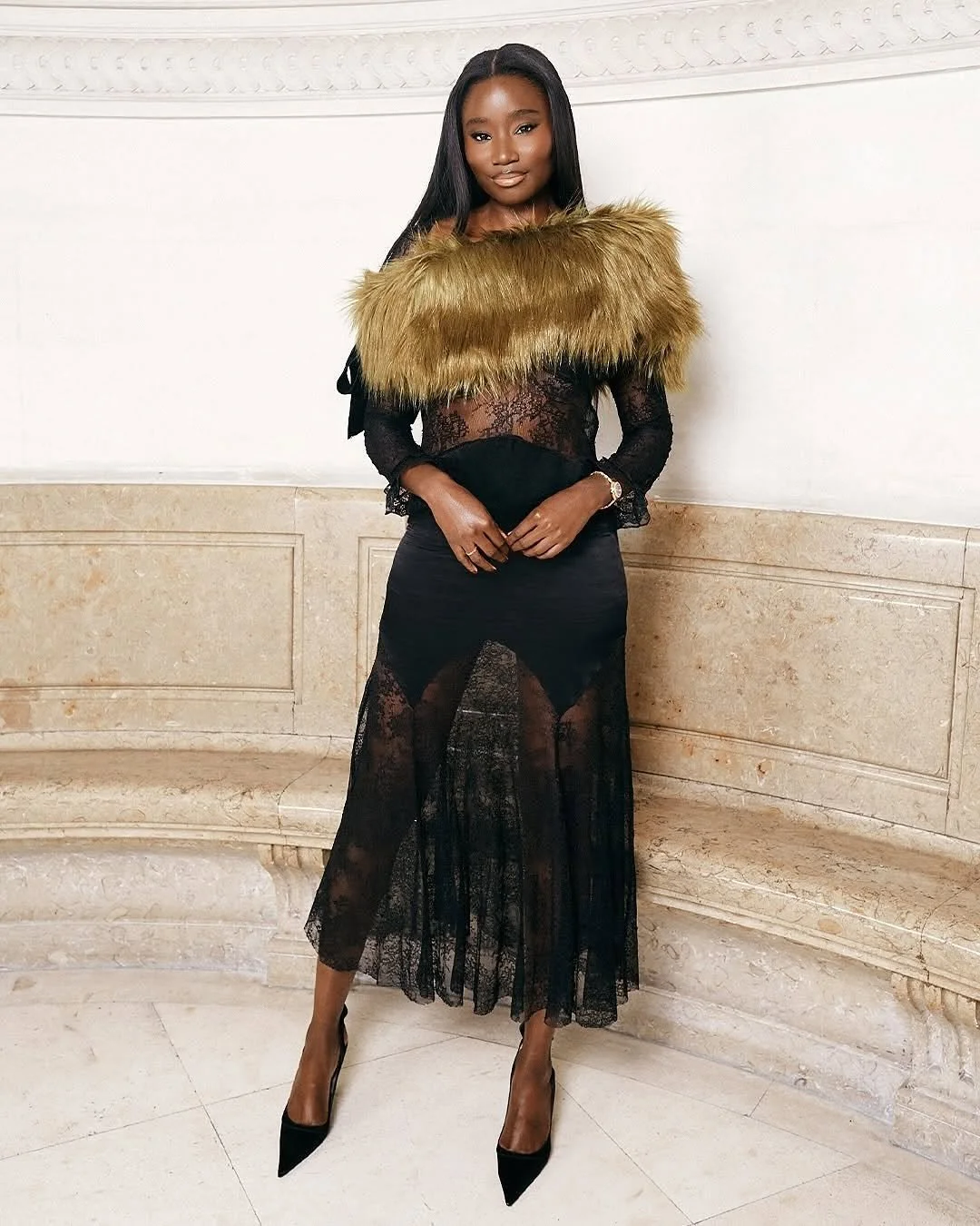 Une femme élégante portant une robe longue en dentelle noire avec une fourrure or foncé sur les épaules, posant dans un cadre intérieur sophistiqué.
