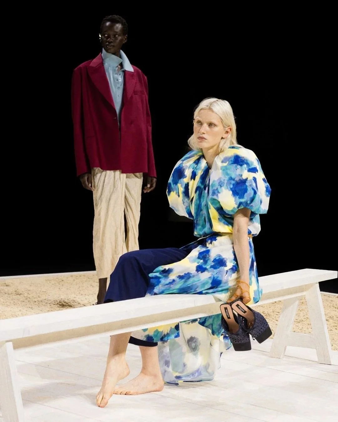 Deux femmes, l'une assise sur un banc, l'autre debout derrière. La femme assise porte une robe fluide à motifs bleus et jaunes, avec des chaussures à talons noires à sequins. La femme debout porte une veste rouge, une chemise bleue, et un pantalon beige. Le fond est noir, et le sol est de la couleur sable.