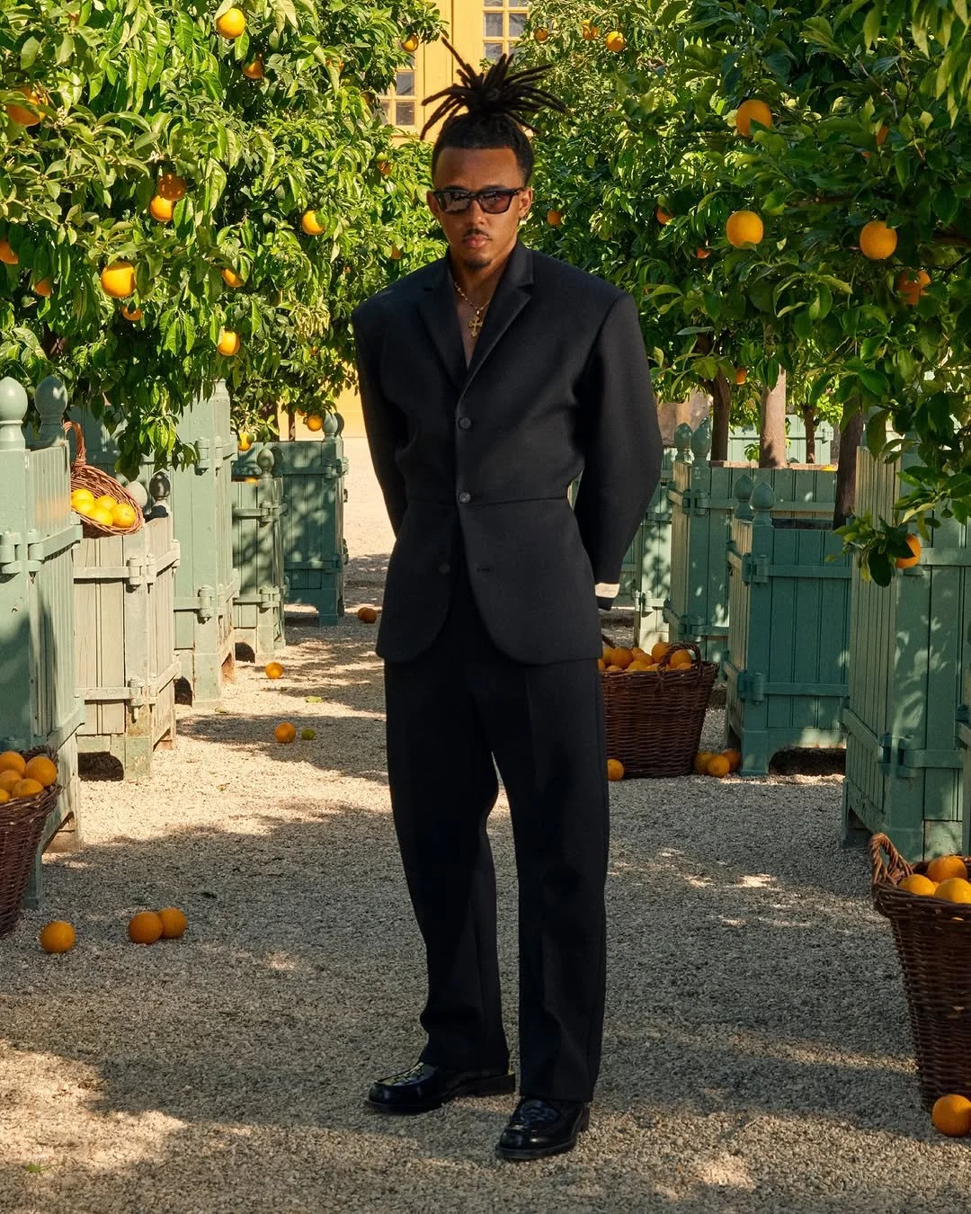 Un homme en costume noir debout dans un verger d'orangers, portant des lunettes de soleil et une chaîne en or.