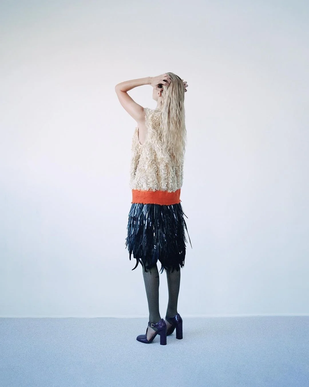 Une femme portant une robe sans manches en laine beige, une jupe en plumes noires, des collants gris et des chaussures à talons violettes, se tient debout contre un fond blanc, la tête tournée de côté avec une main dans les cheveux.