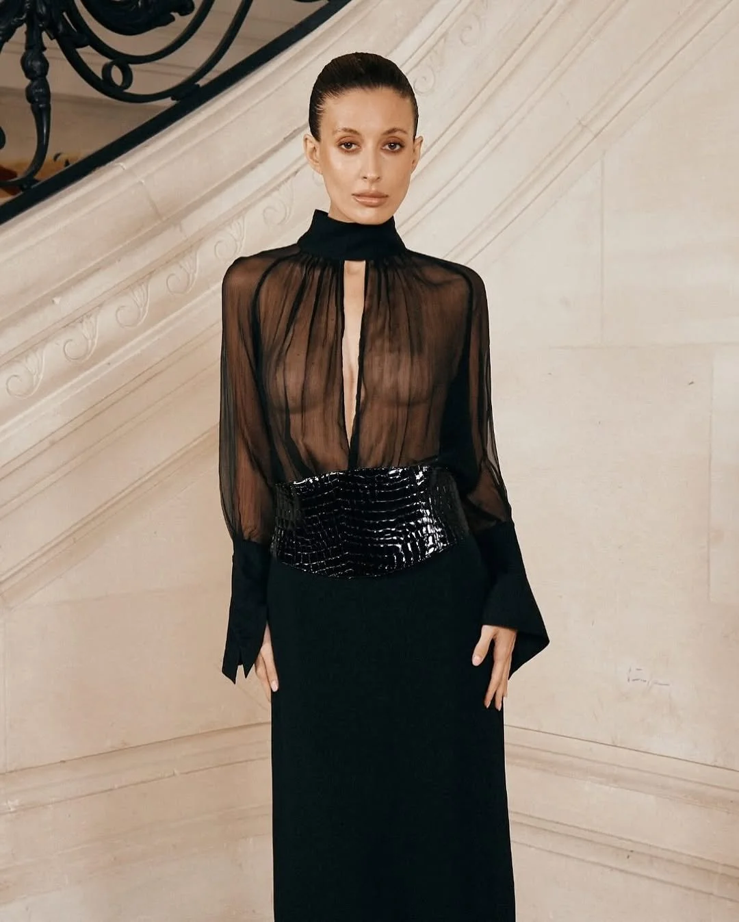 Une femme portant un haut en tissu transparent noir avec un col montant et une jupe longue noire, posant devant un mur en pierre beige avec un escalier en fer forgé en arrière-plan.