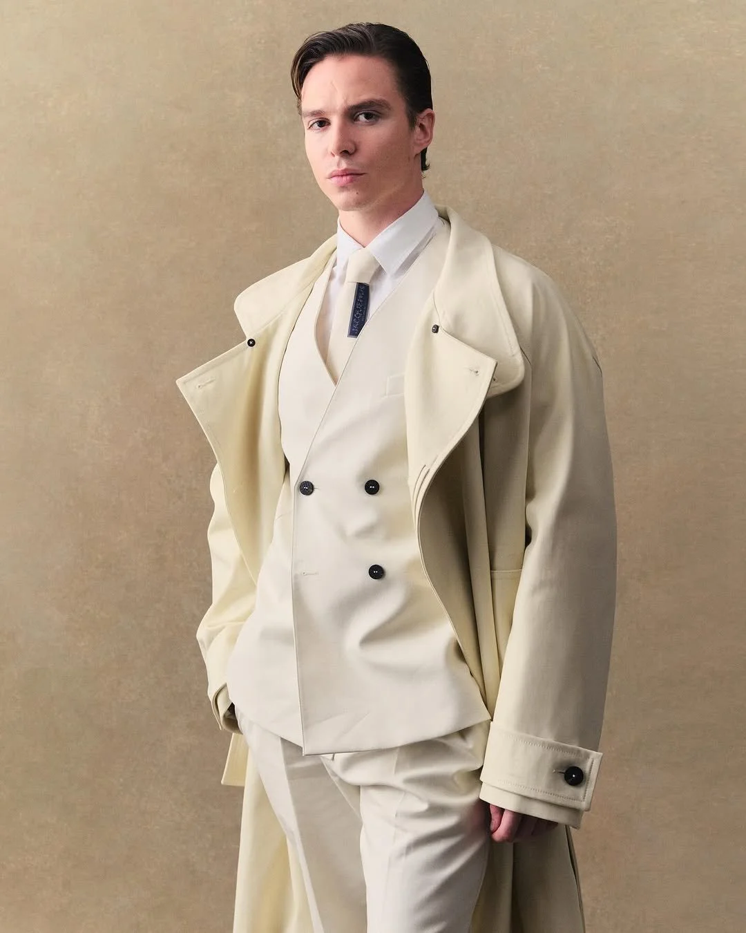 Jeune homme portant un costume élégant beige clair avec une cape en laine de même couleur, debout contre un fond beige, posant avec une expression sérieuse.