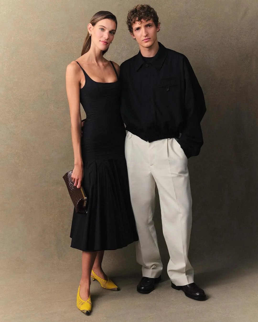Un homme et une femme posant ensemble contre un fond beige. La femme porte une robe noire élégante avec des talons jaunes, et l'homme porte un haut noir avec un pantalon clair.