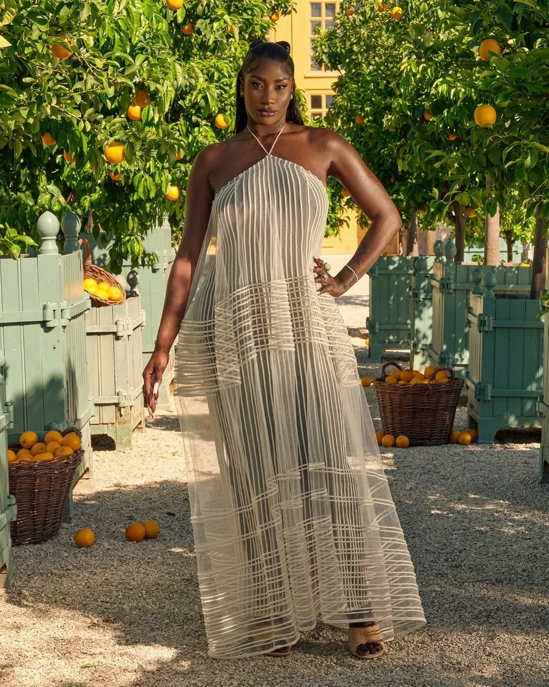 Une femme en robe longue, beige avec des rayures, posant dans un verger d'orangers, avec des paniers d'oranges autour.