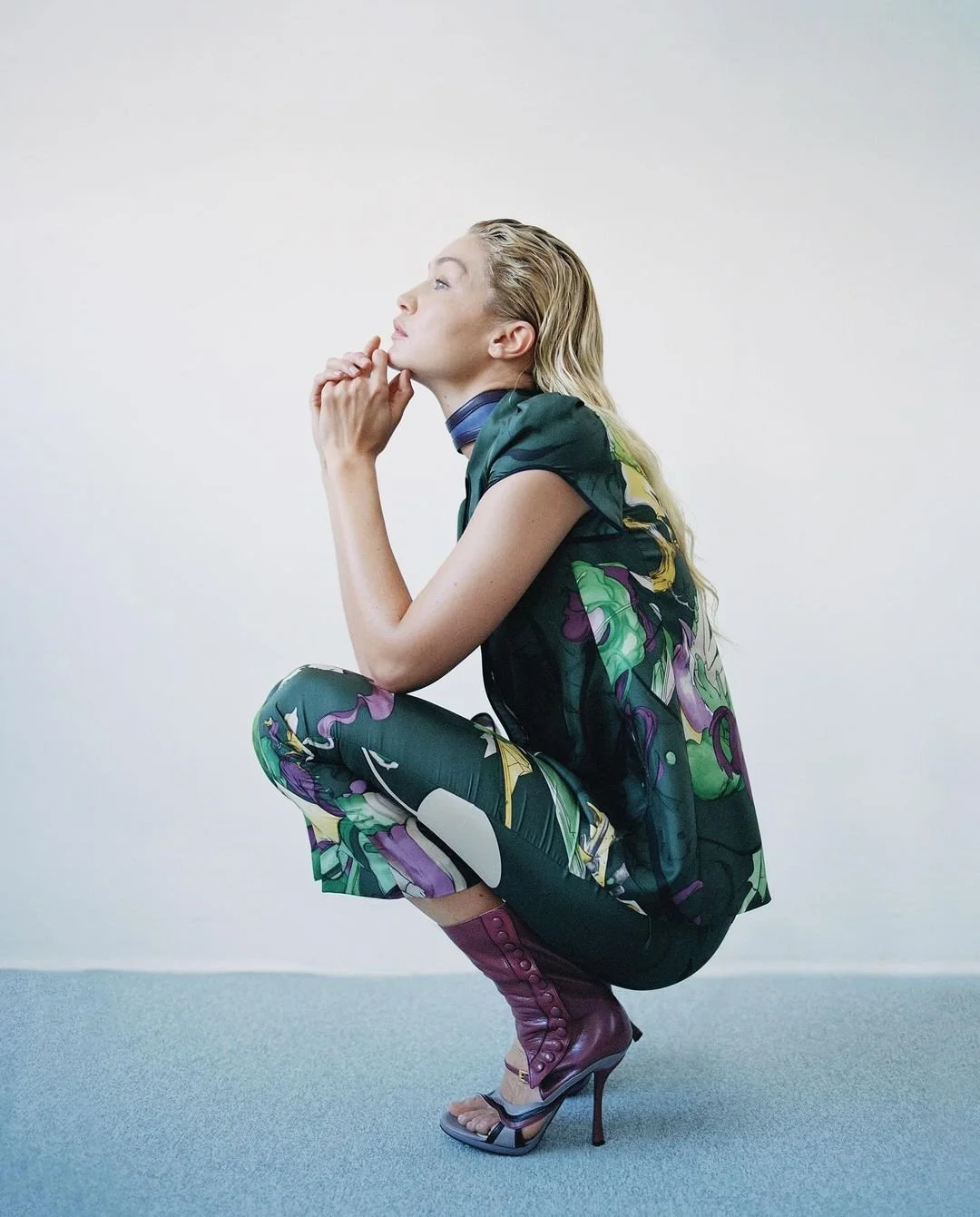 Femme en vêtement à motifs floraux, portant des bottes à talons, en position accroupie, le menton posé sur ses mains, regardant vers le haut, dans une pièce vide.