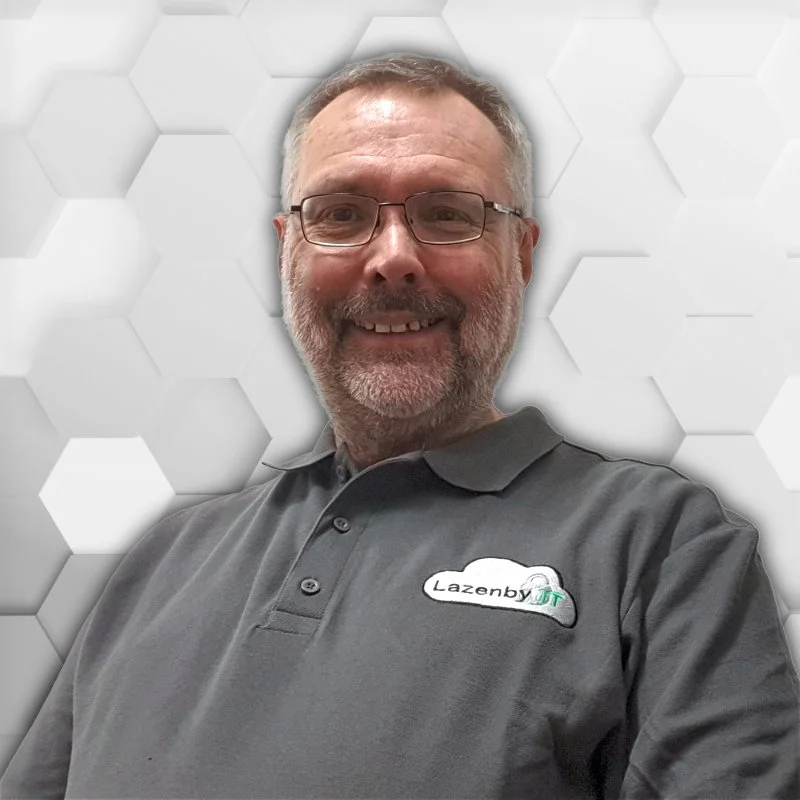 A man with glasses and gray hair smiling, wearing a dark gray polo shirt with a Lazenby IT logo, in front of a white hexagonal patterned background.