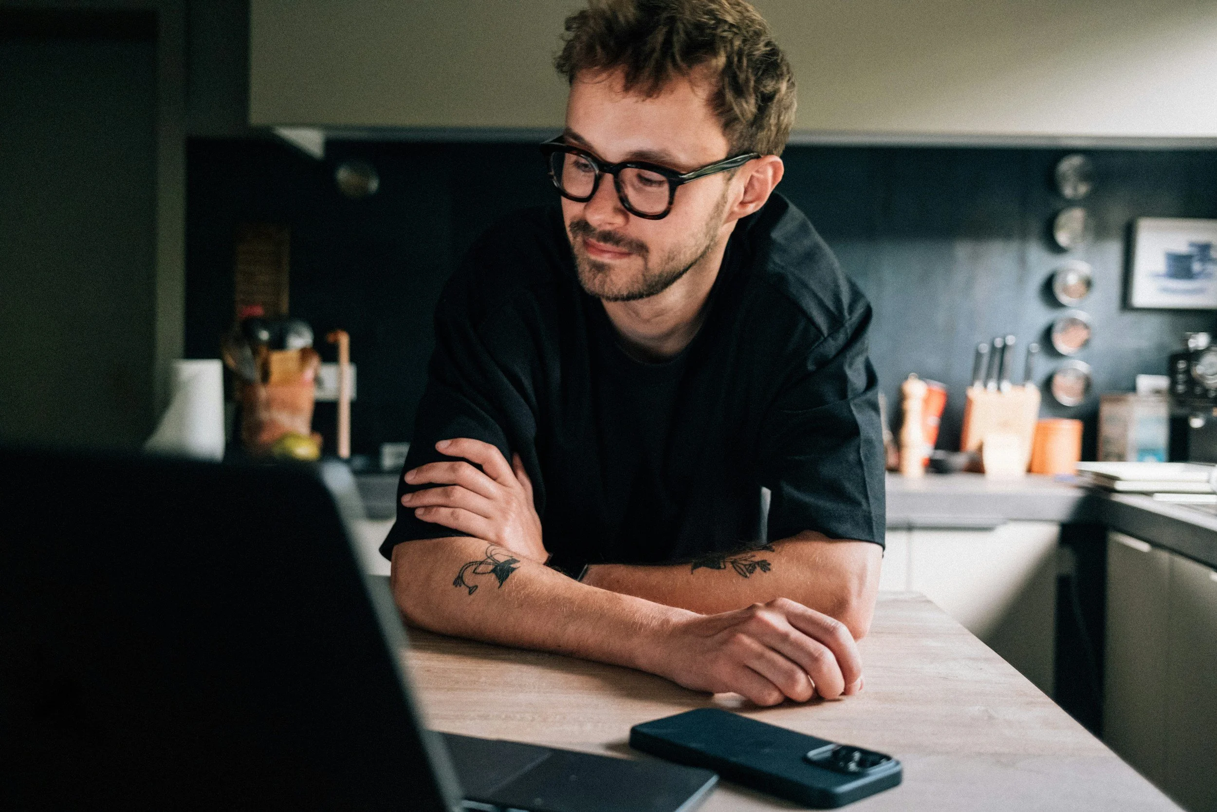 A man with glasses and tattoos sitting at a kitchen counter, looking at a laptop, with a smartphone on the table.