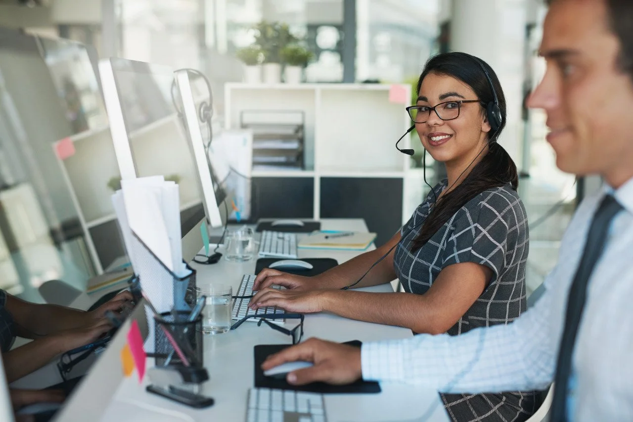 A woman with glasses and a headset smiling at a computer in an office, with a man sitting nearby.