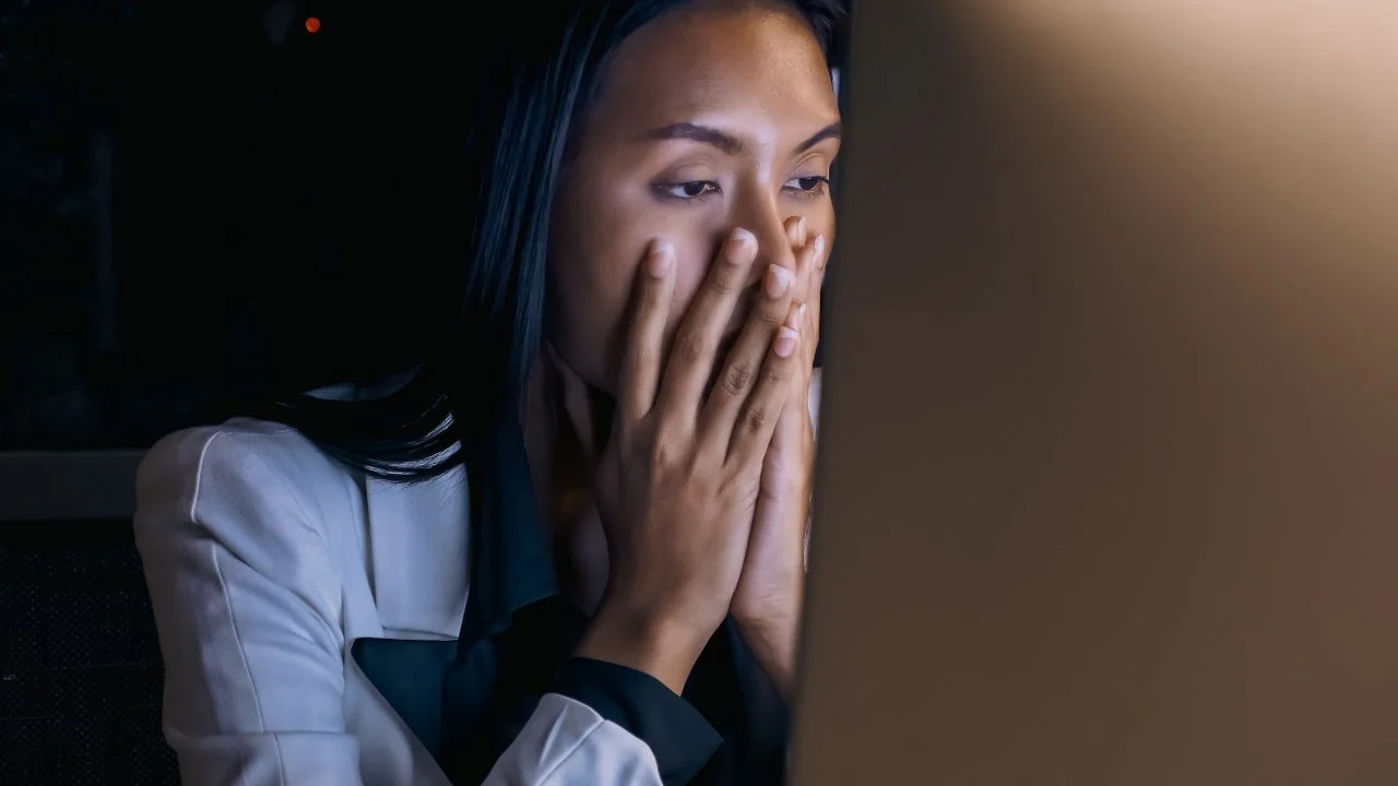 Woman with long dark hair looking at a screen with a concerned expression, hands covering her mouth.