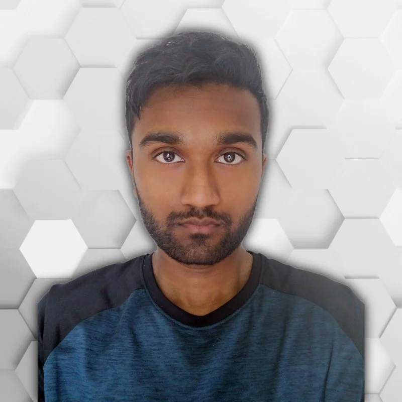 A young man with dark hair, a beard, and brown eyes looking at the camera, wearing a blue and black shirt, against a white geometric honeycomb-patterned background.
