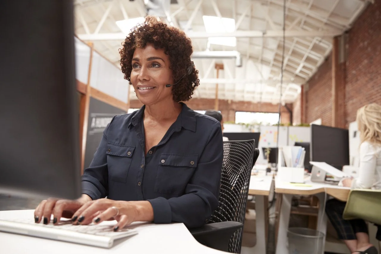 A woman wearing a headset working at a computer in an open-plan office with exposed brick walls and high ceilings.