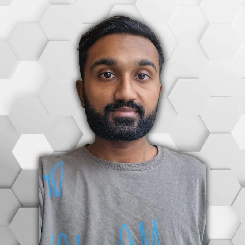 A man with dark hair and beard taking a selfie in front of a white hexagonal textured wall. He is wearing a light gray t-shirt with blue text.